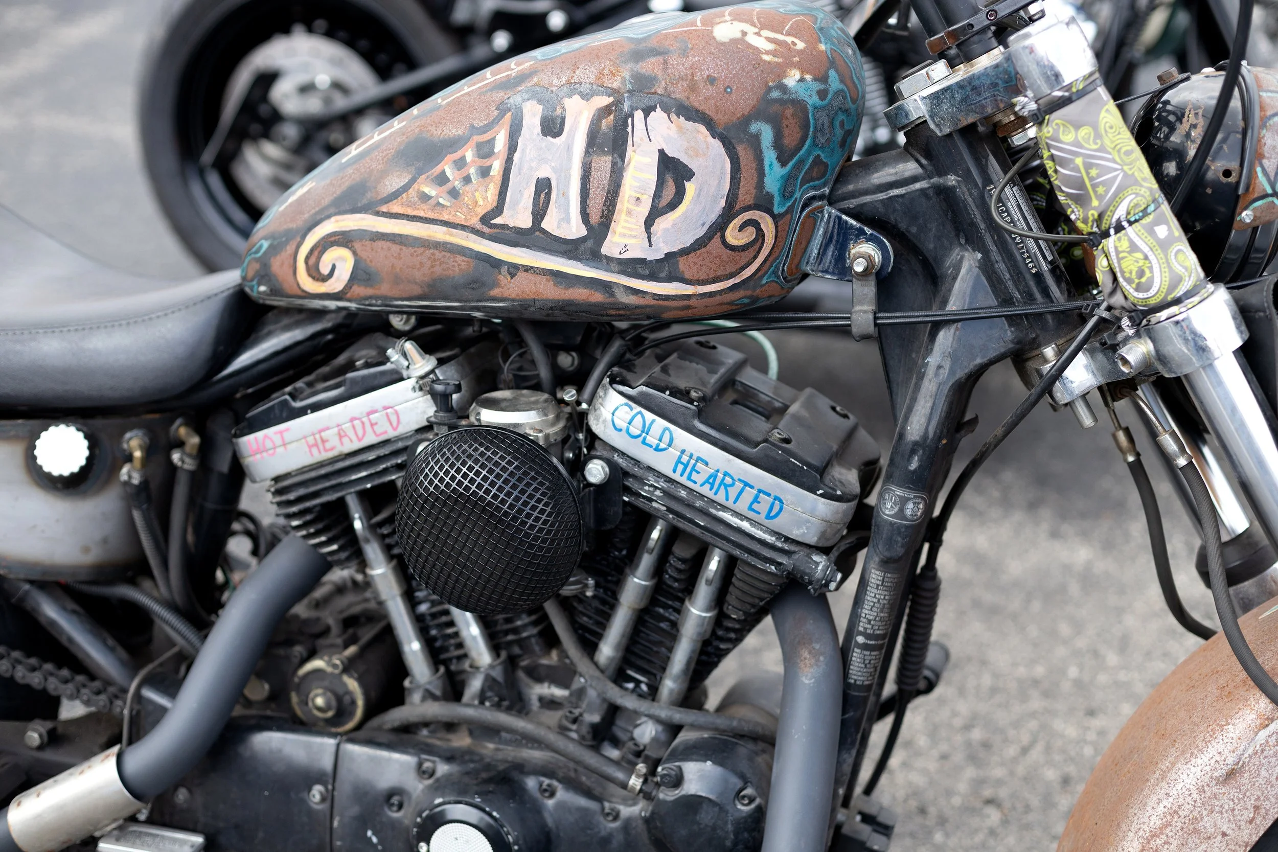 Close-up of a motorcycle with a rusted metal fuel tank featuring painted designs and words, surrounded by various engine parts and cables.