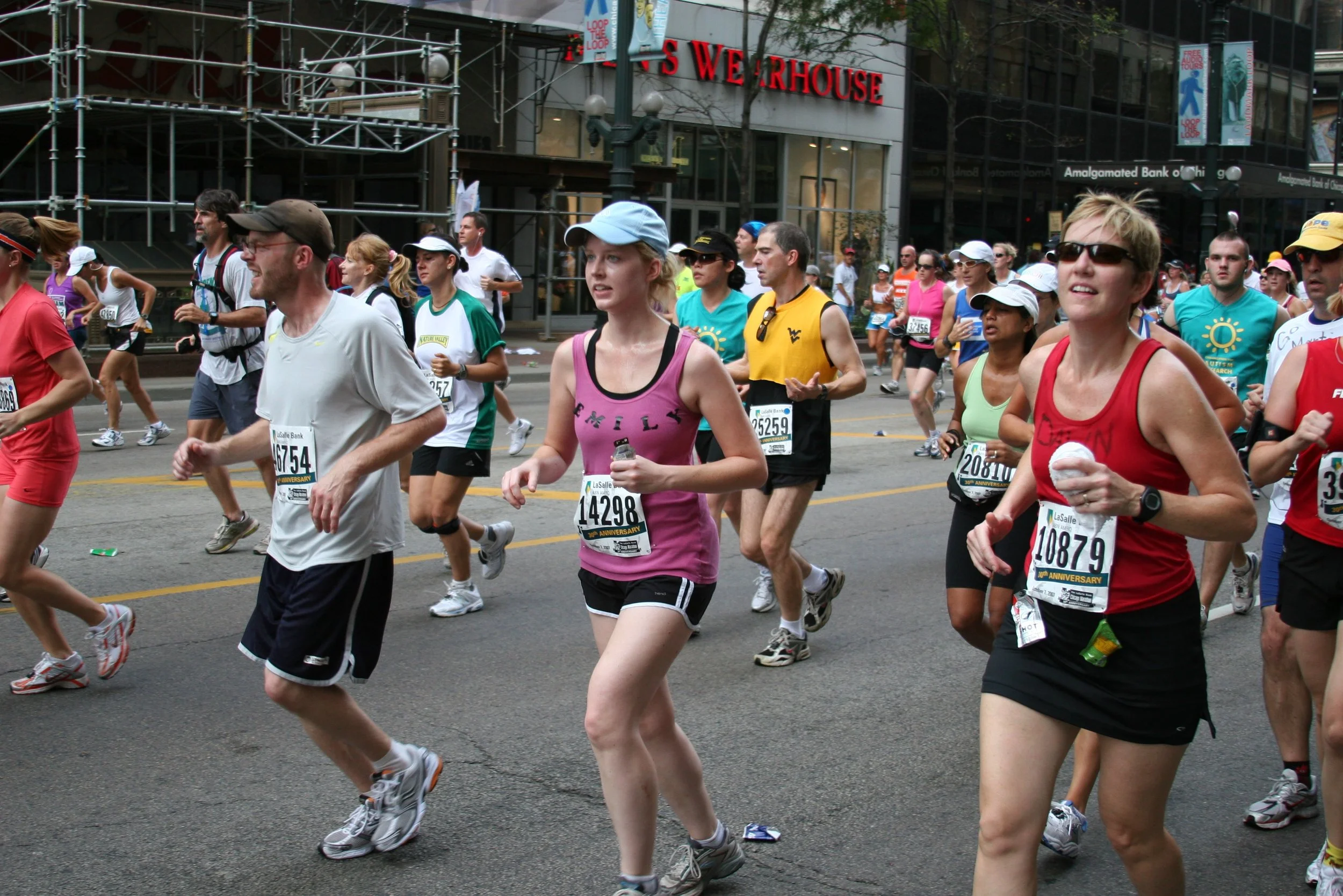 A group of marathon runners participating in a race on a city street, with buildings and storefronts, including a sign for 'S WAREHOUSE,' in the background.