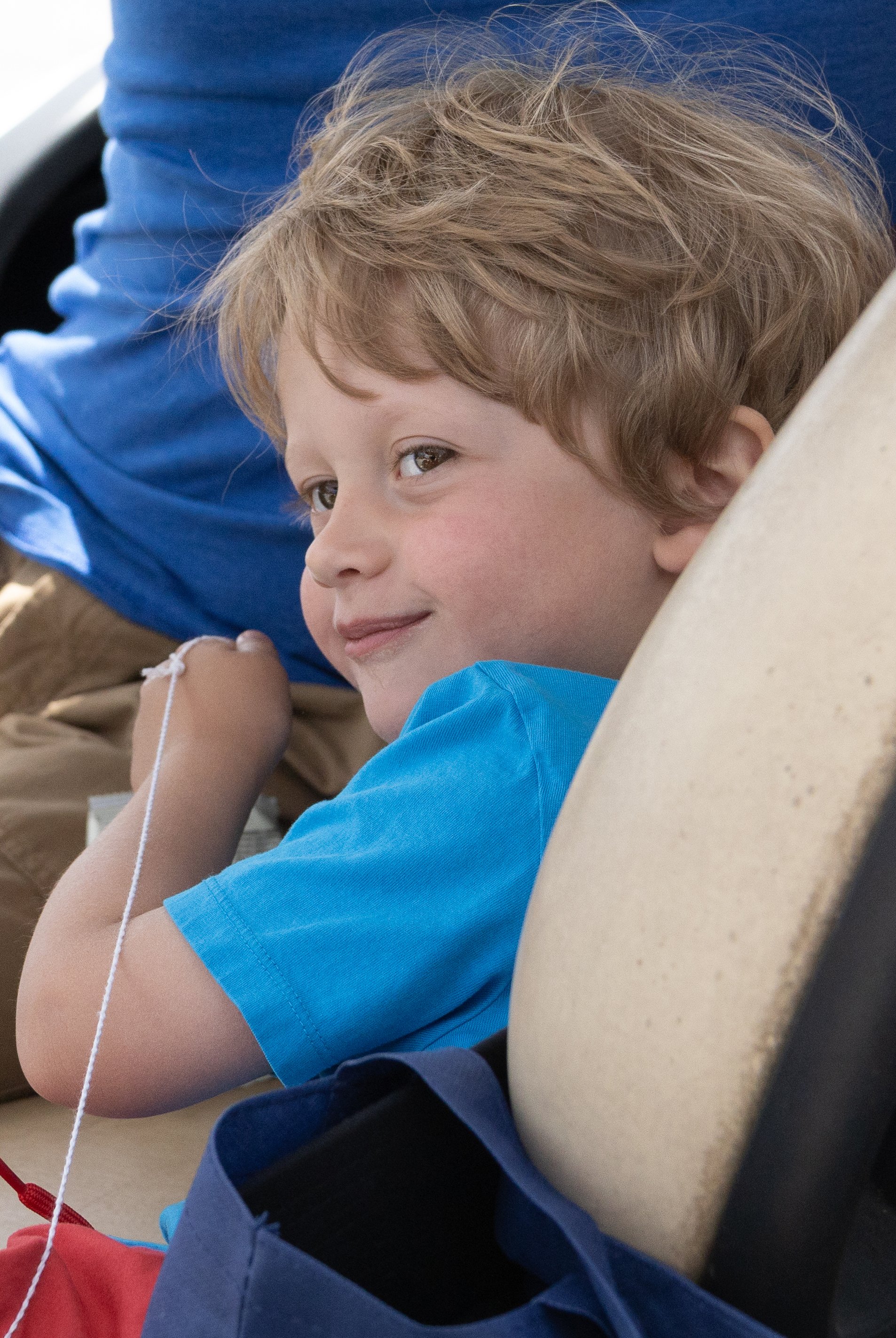 A young boy with light brown, wavy hair, wearing a bright blue shirt, lying on a beige surface, possibly on a plane or hospital bed, with a slight smile on his face. He is holding an IV line attached to his arm, with various bags and blue fabric near