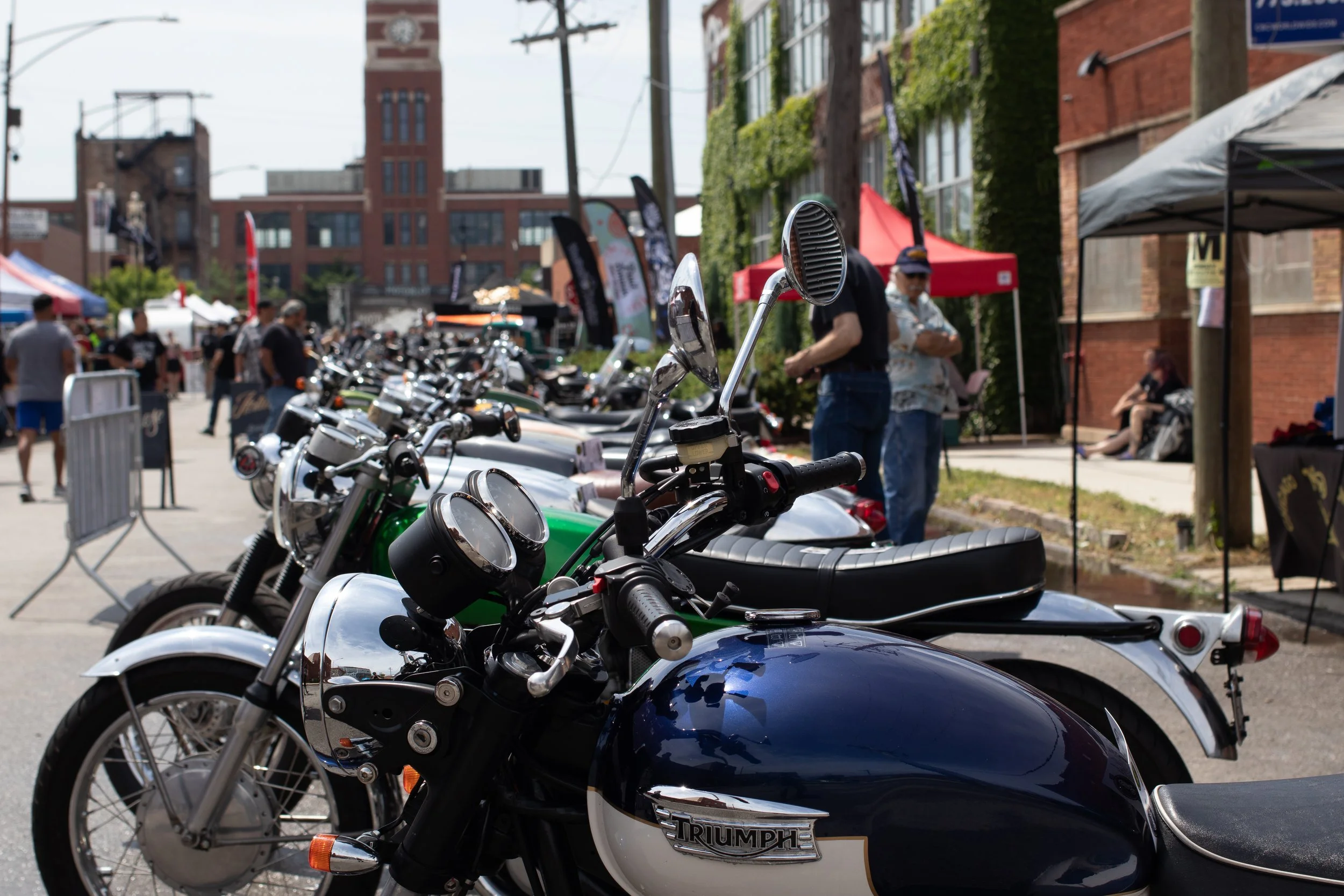 Close-up of several parked Triumph motorcycles at an outdoor event with tents and people in the background.