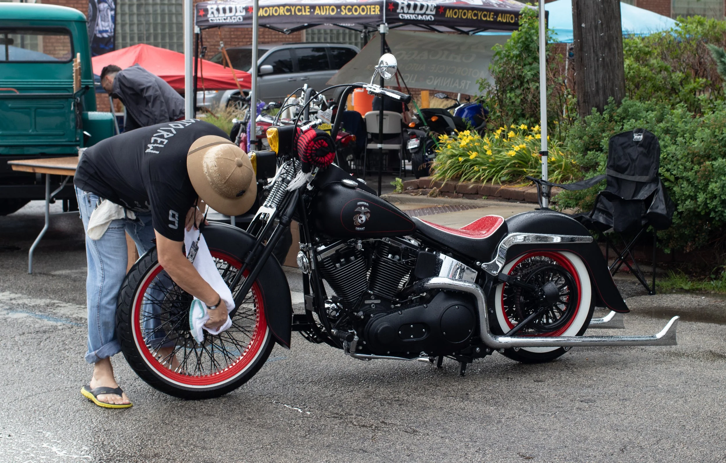 A man wearing a cowboy hat, black t-shirt, and flip-flops is cleaning or inspecting a black motorcycle with red accents and a decal of a bulldog on the side, parked in a wet parking lot. Behind him, there is a canopy with motorcycle and auto parts fo