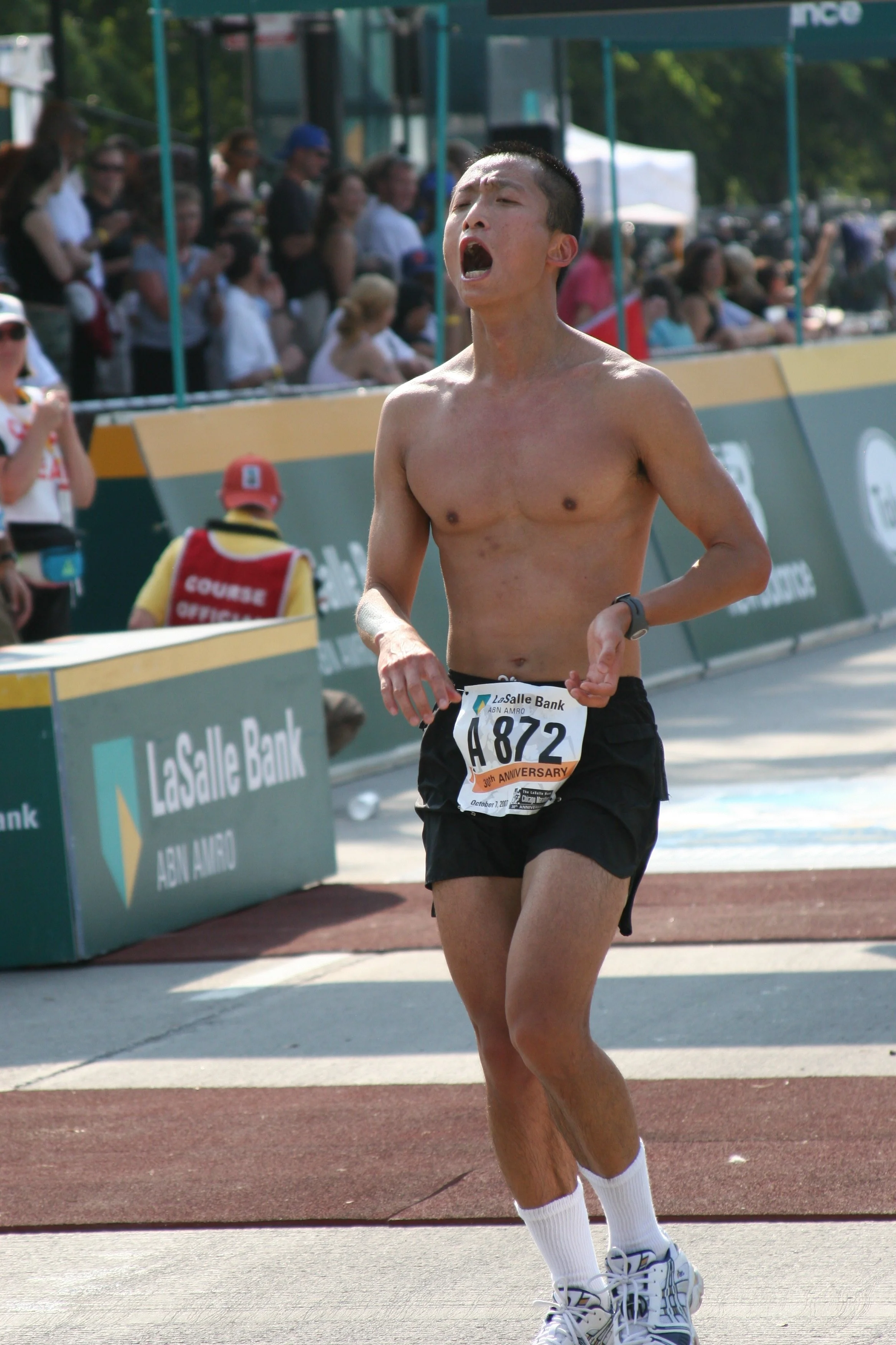 A male runner in black shorts and white sneakers running on a track with an open mouth, shirtless, and wearing a watch. Spectators and event staff are visible in the background, along with various banners and signs, including one for LaSalle Bank.