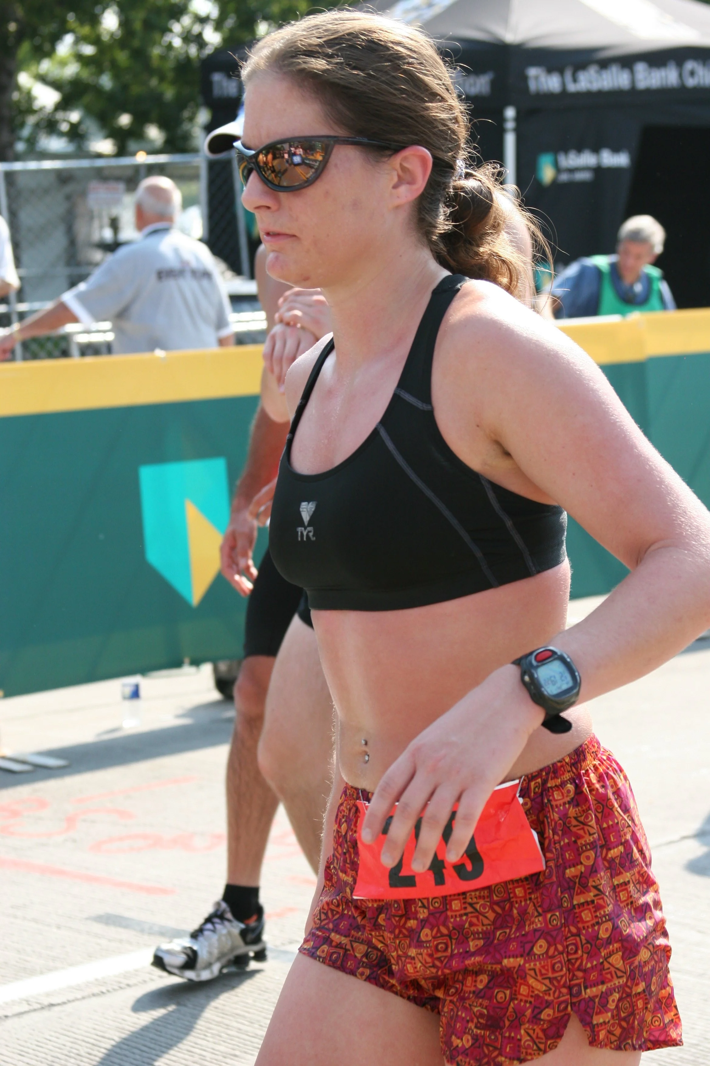 A female runner wearing sunglasses, a black sports tank top, and colorful patterned shorts, with a sports watch on her wrist, participating in a race or marathon outdoors.