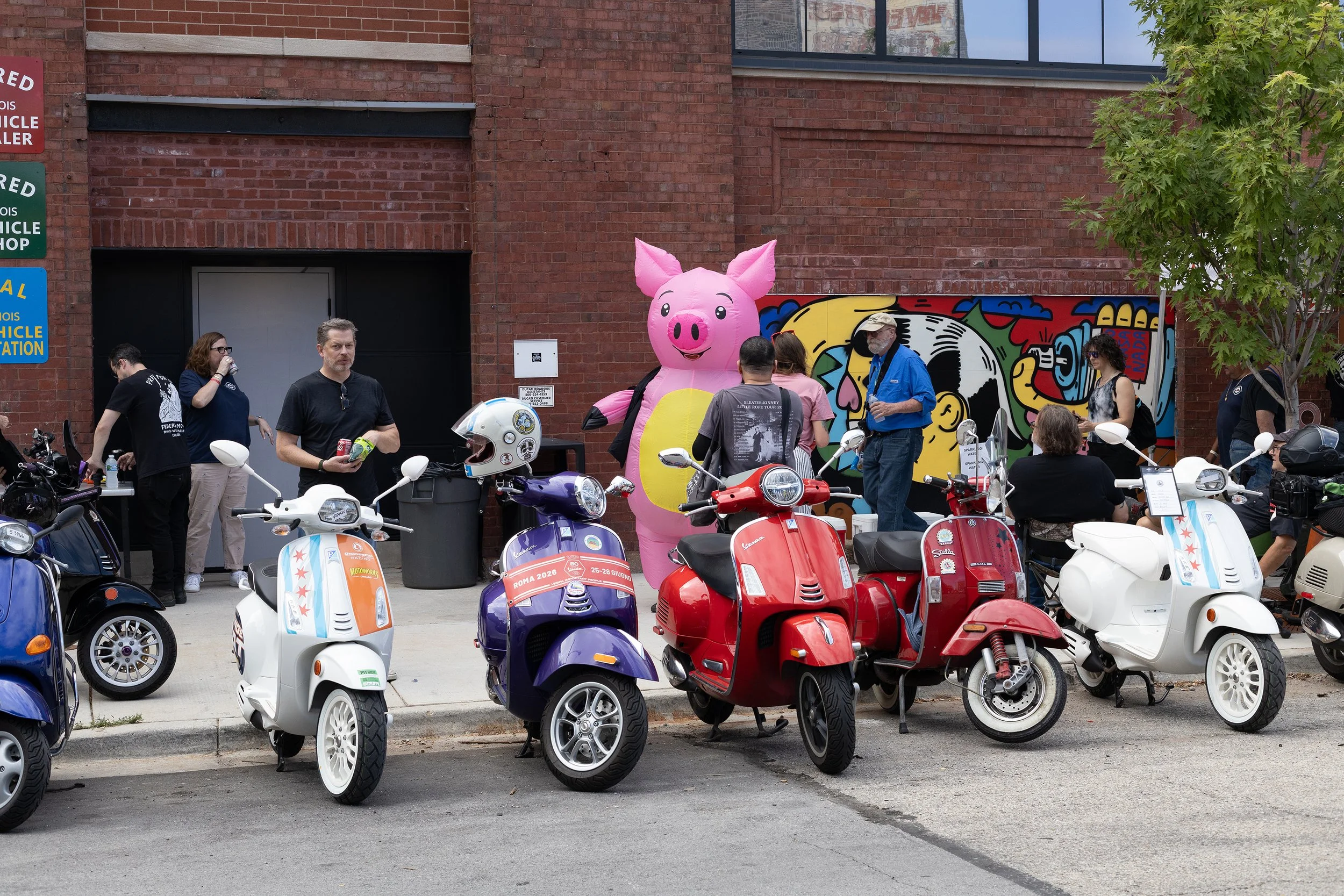 A group of people gathered outside a brick building with colorful graffiti art in the background. Multiple scooters are parked in the foreground, and a large inflatable pink pig is visible behind the people.