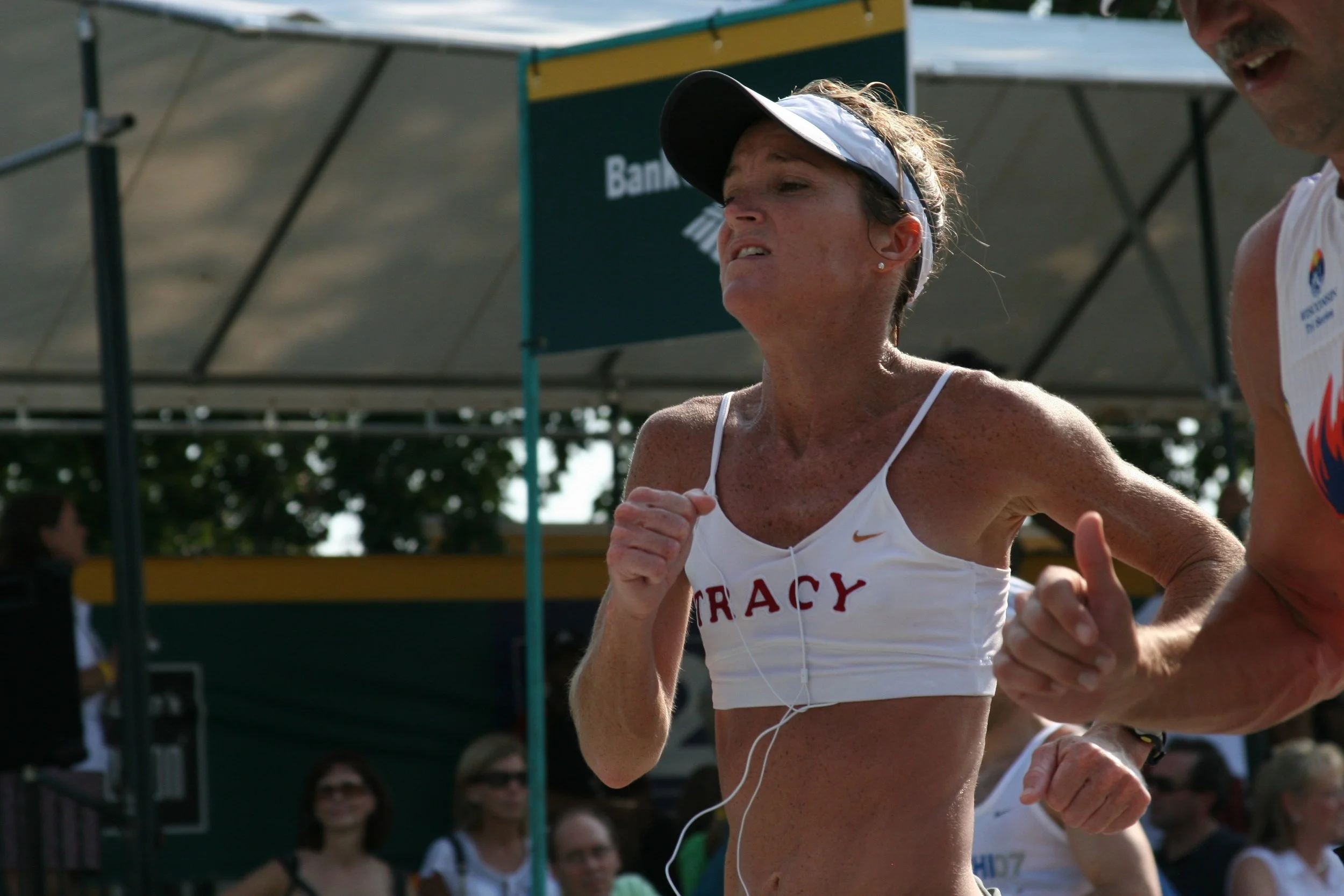 A female runner with freckles wearing a white tank top labeled 'TRACK', a white visor, and earphones, running in a race with a man beside her. The background shows a green banner and spectators.