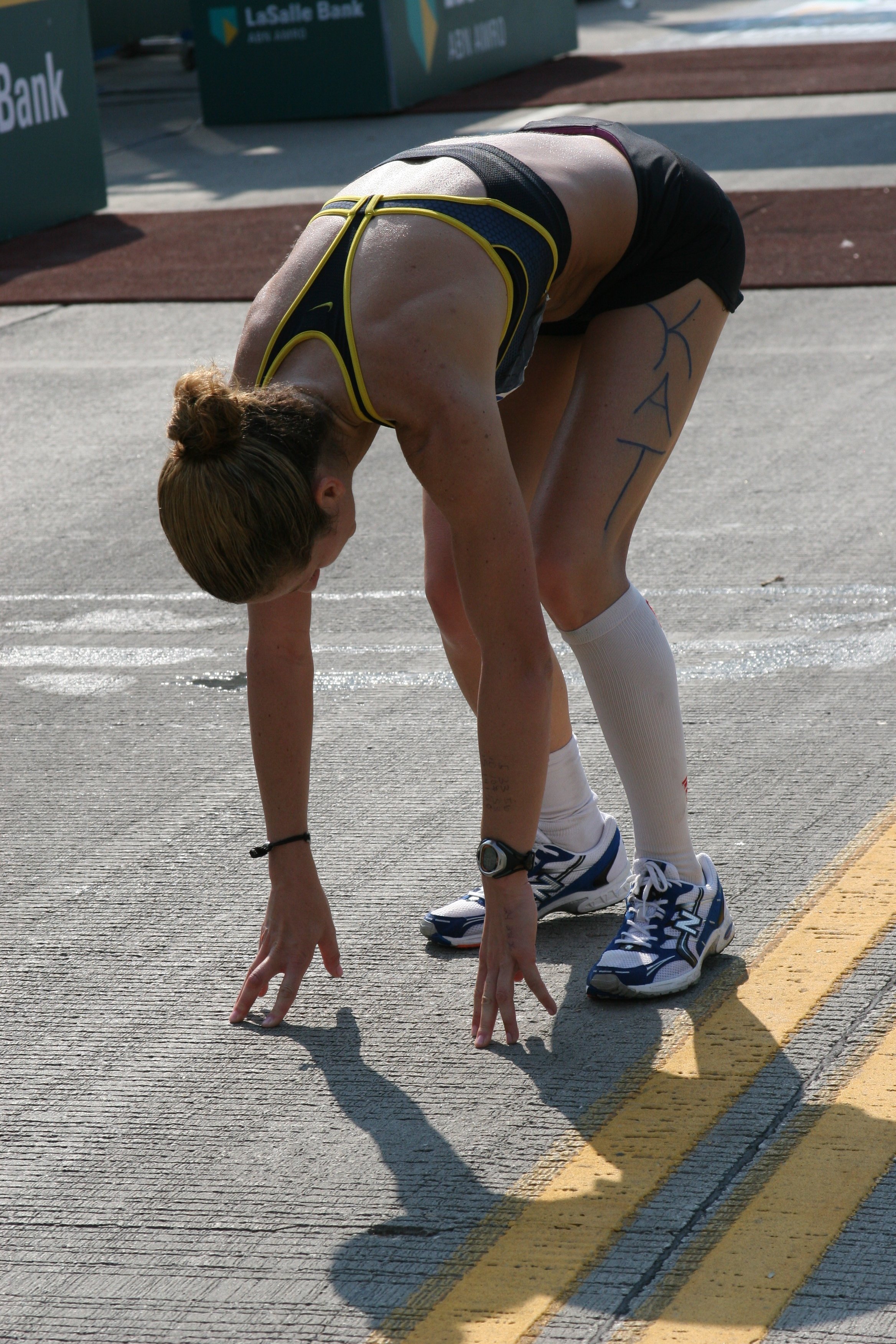 A woman in athletic attire prepares for a running race at the starting line on a city street. She is bent over with her hands and feet on the ground, wearing sneakers, shorts, and a sports top, with her hair in a bun and the word "KAY" written on her