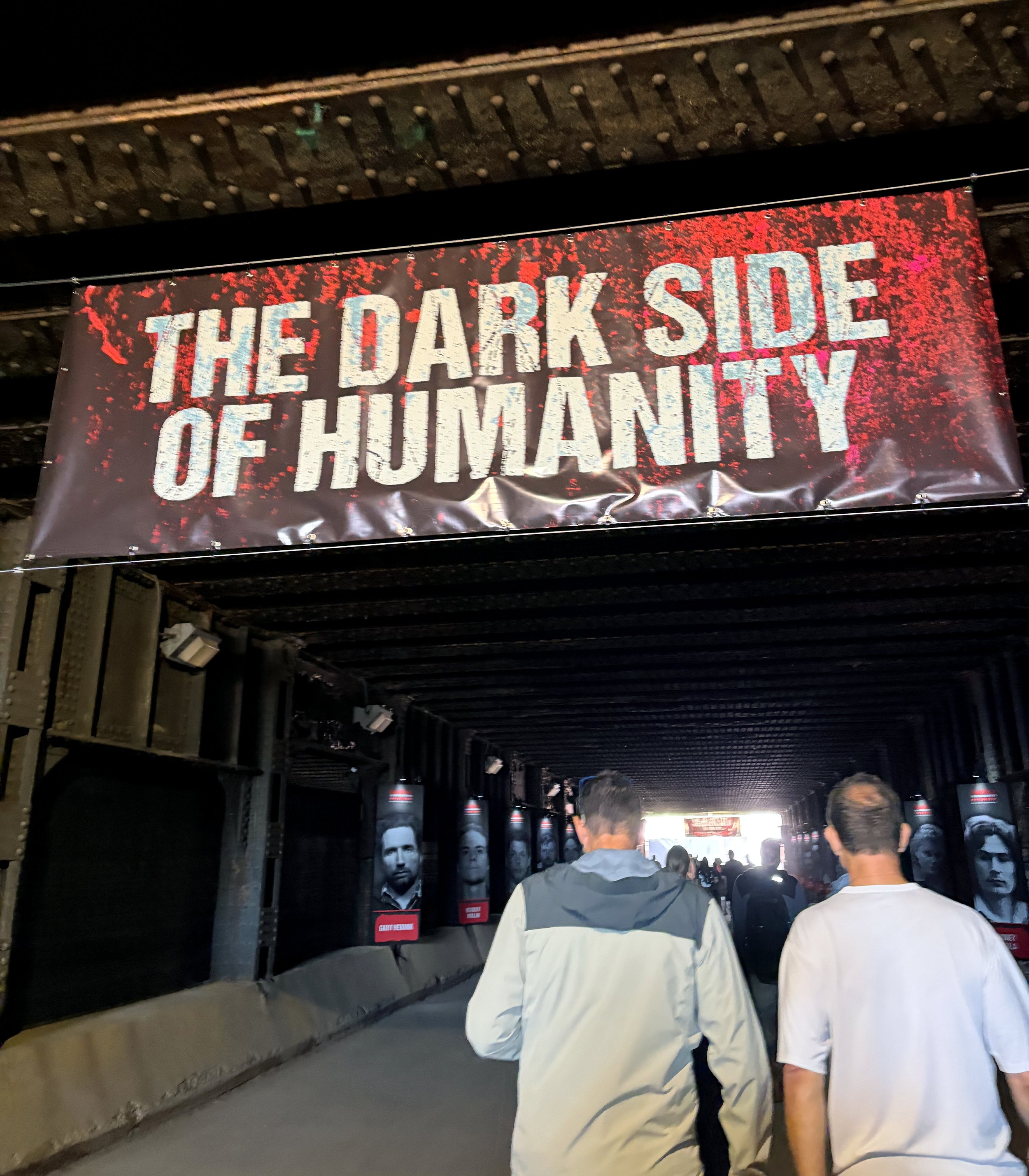 A dark hallway with a large banner hanging that reads 'The Dark Side of Humanity' in bold white letters with a red and black splattered background. People are walking under the banner, and portraits of men's faces are displayed on either side of the 