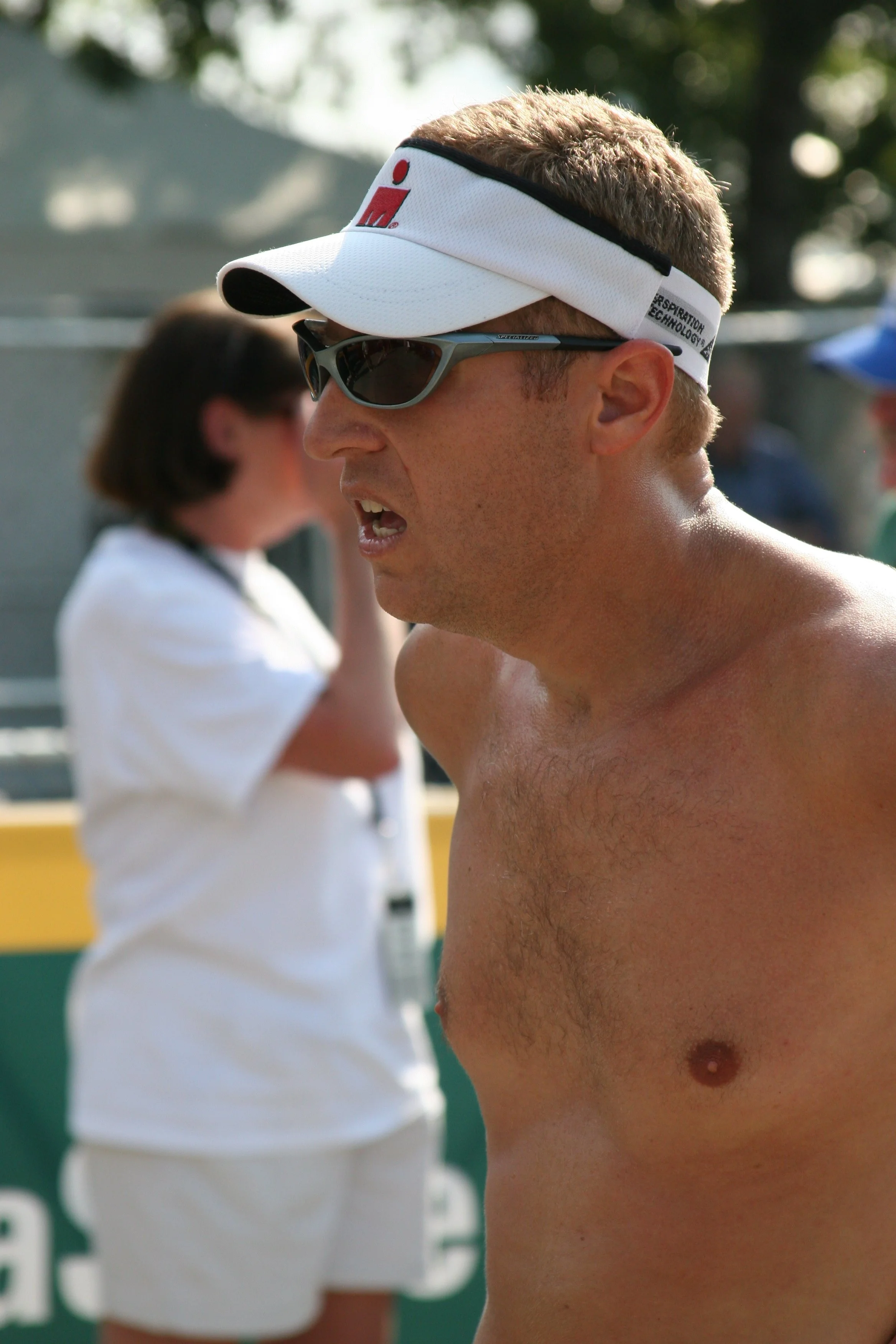 Close-up of a shirtless man wearing sunglasses and a white visor cap, with a sunny outdoor background. A woman in a white shirt is blurred in the background.