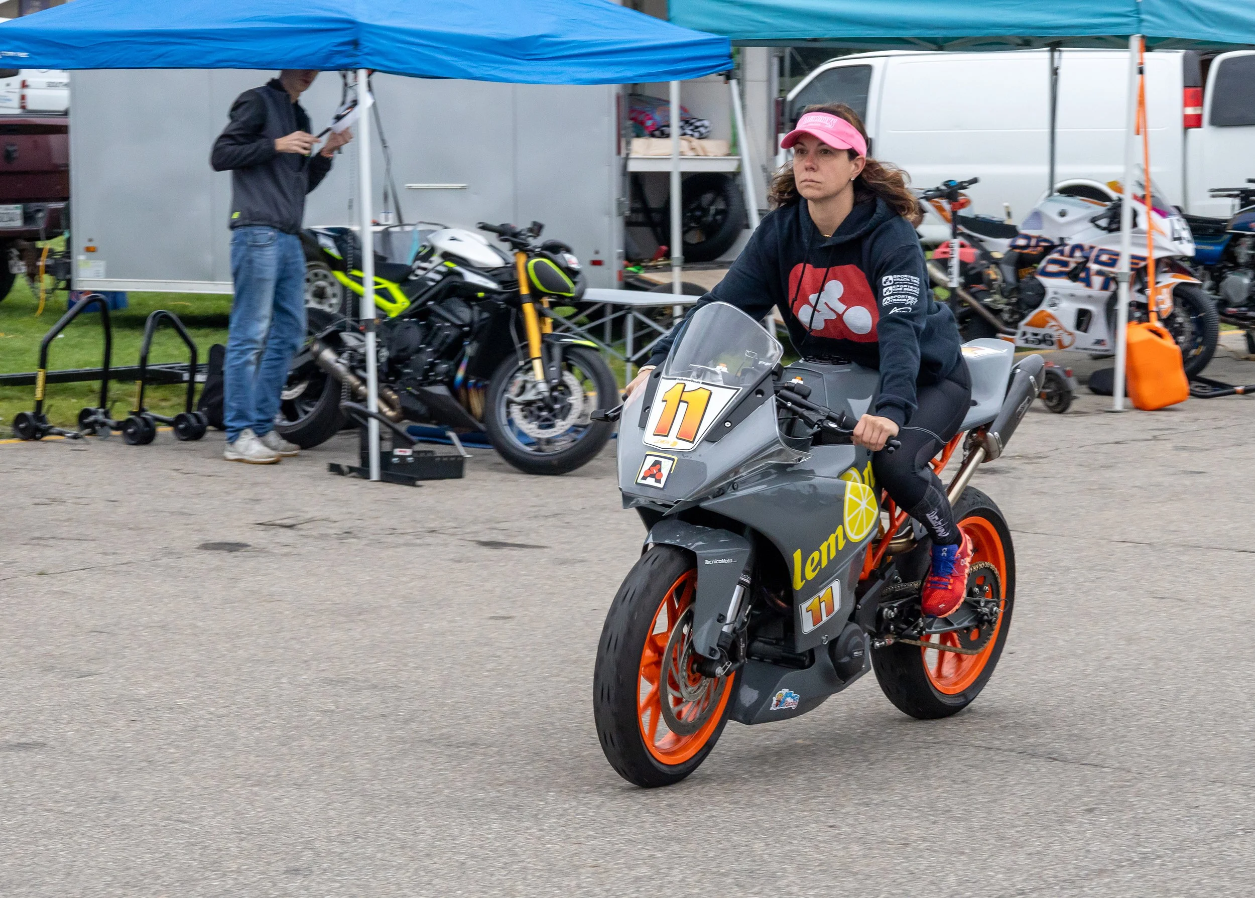 A woman riding a gray and orange electric motorcycle with the number 11 on it, wearing a pink visor hat and a black hoodie, at a racing event with tents and motorcycles in the background.