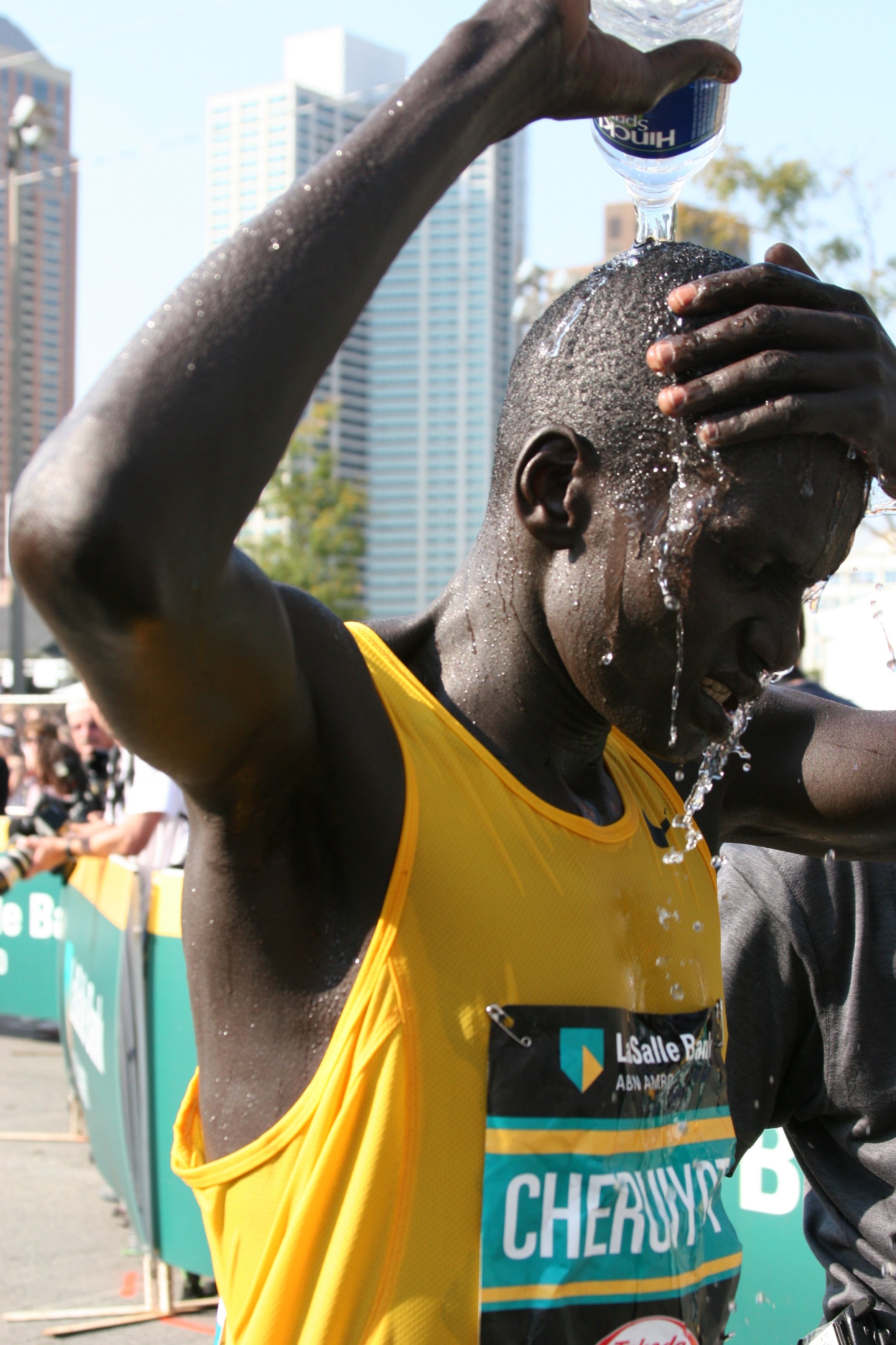 A runner with dark skin and a shaved head tilting her head back, pouring water over her head from a plastic bottle during a race, wearing a yellow running shirt and a race bib with the name 'Cheruk' on it.