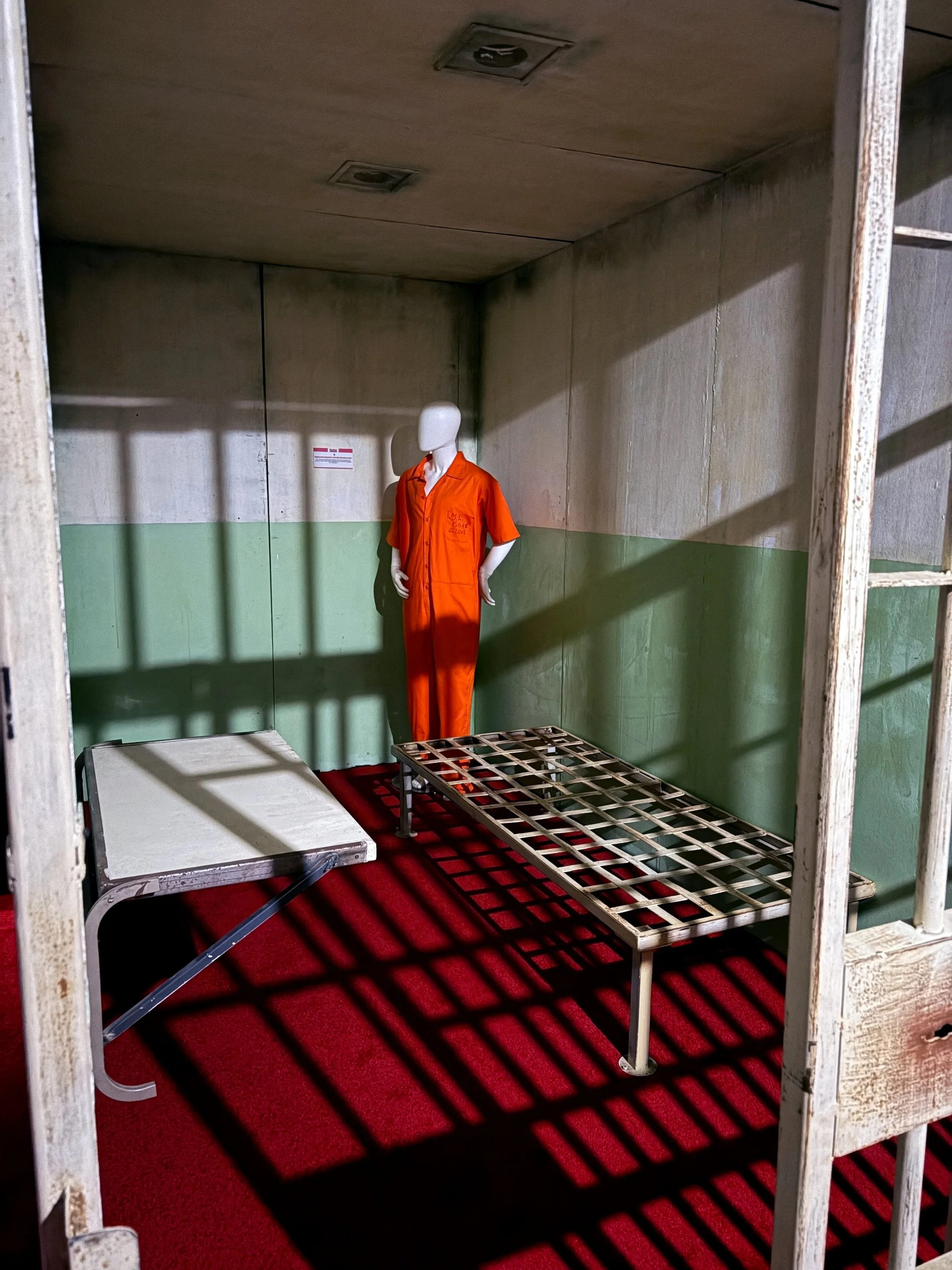 Empty prison cell with a mannequin wearing an orange jumpsuit, a metal bed frame, and a small white table, with red carpet flooring and shadowed walls.