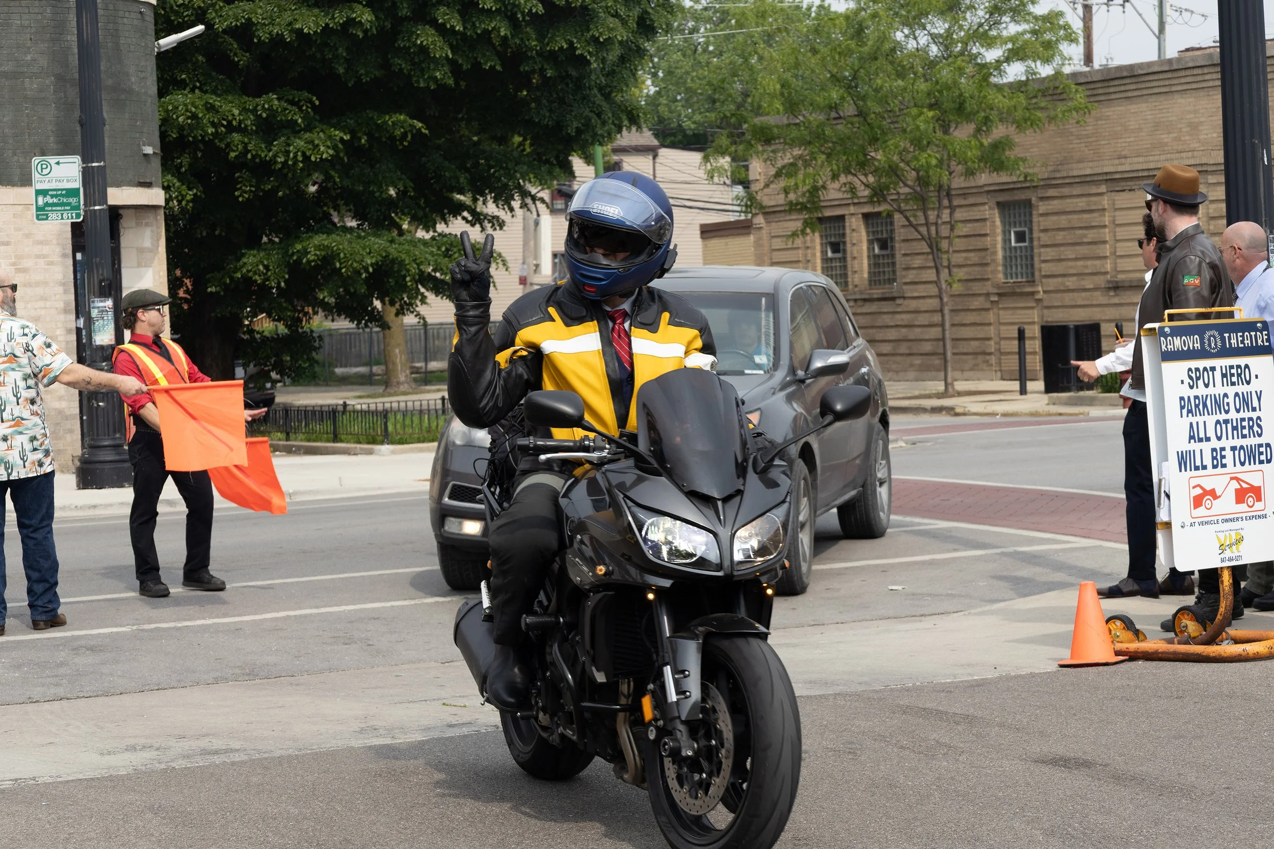 A motorcyclist in a helmet and jacket making a peace sign while stopped at an intersection. There are several people and a sign nearby, with trees and buildings in the background.