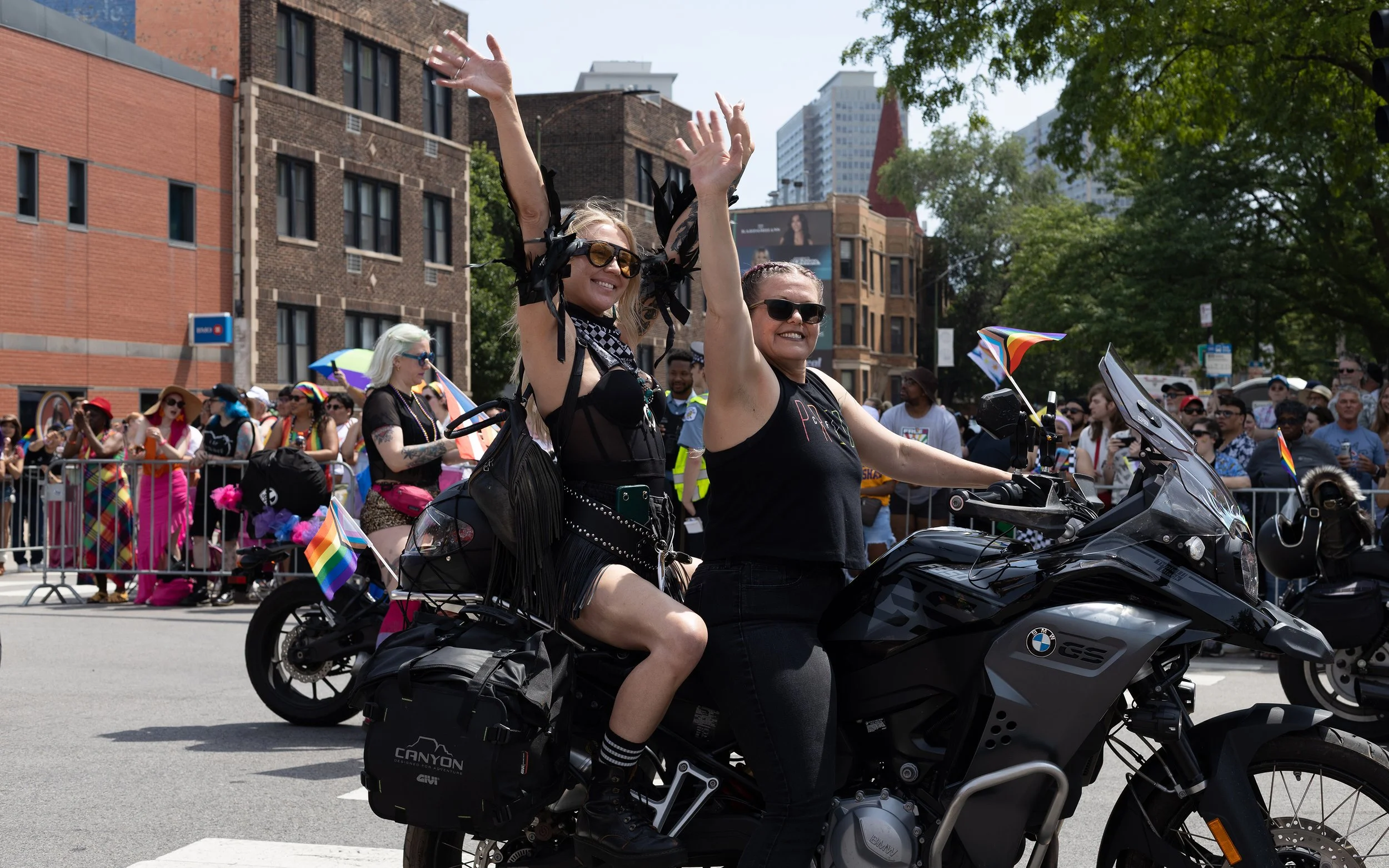 Two women riding a motorcycle during a parade or festival, surrounded by a crowd of people, some holding rainbow flags, with city buildings and trees in the background.