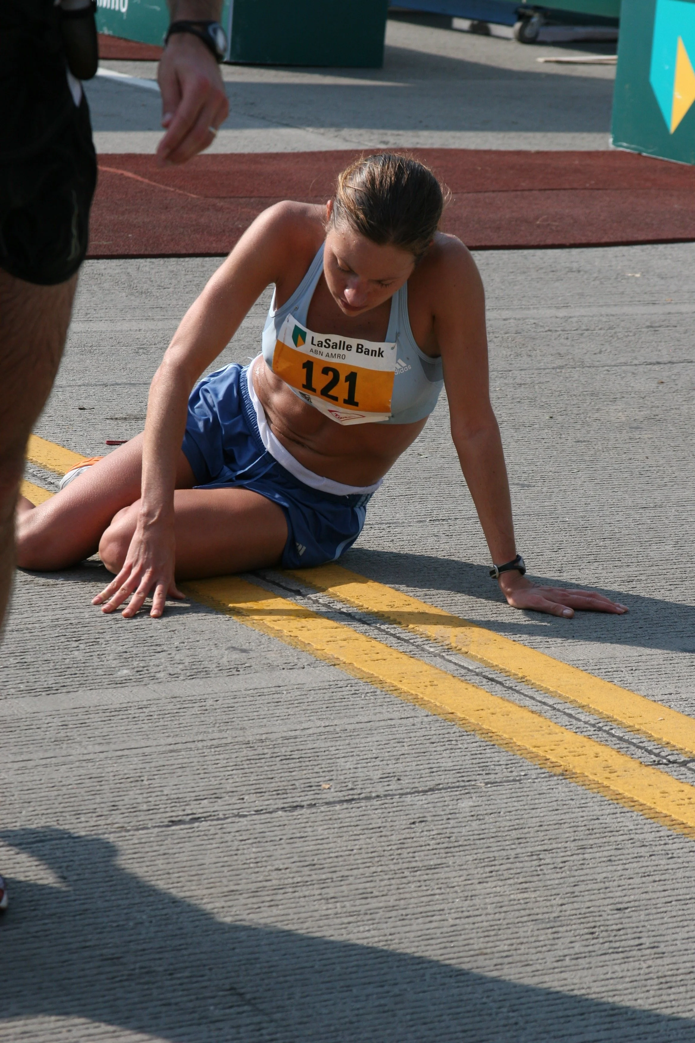 A female runner resting on the ground after a race, wearing a light blue top and blue shorts, with a race bib number 121 and sponsor LaSalle Bank, on a road with yellow lines and red track surface in the background.