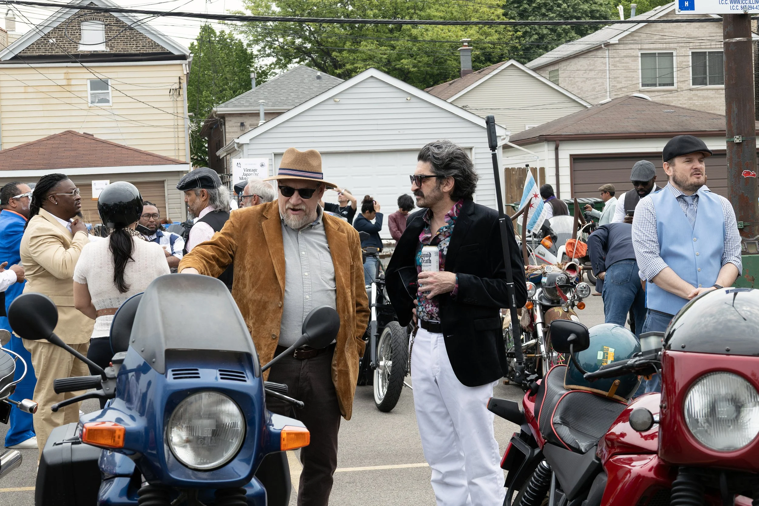 A group of people at an outdoor event in a residential neighborhood, standing around scooters and motorcycles, with three main men in the foreground having a conversation, while others are in the background near houses and trees.