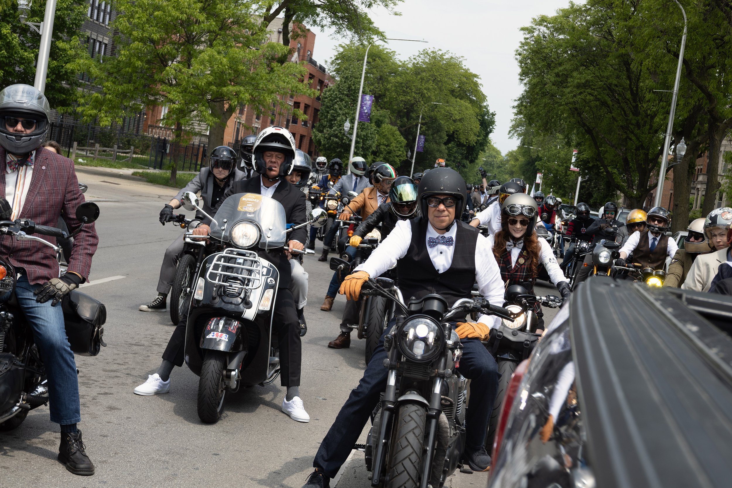 A diverse group of people riding motorcycles and scooters on a city street, with green trees and buildings in the background.