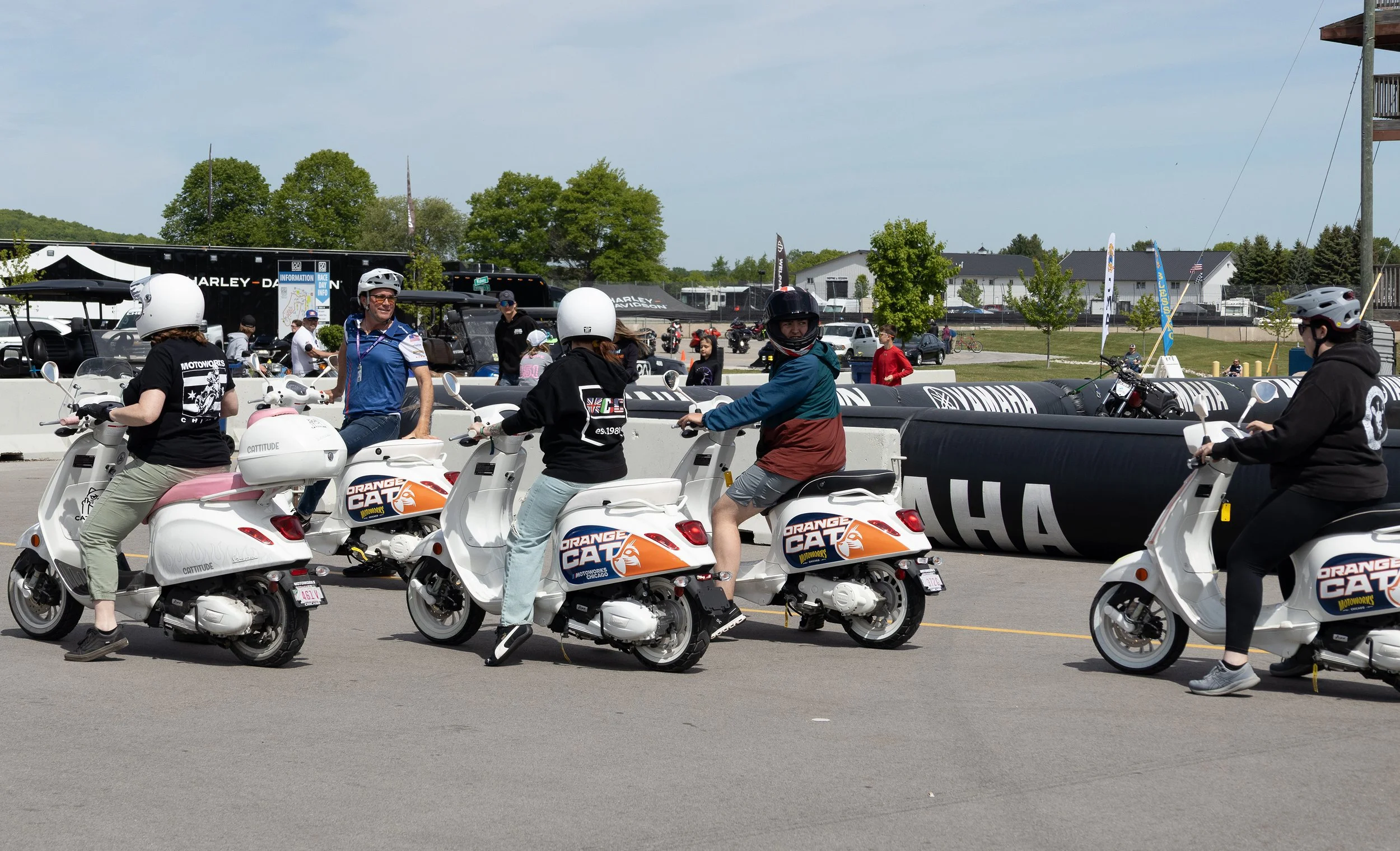 People on white scooters waiting in line at an outdoor event, some wearing helmets, with a Harley-Davidson trailer and trees in the background.