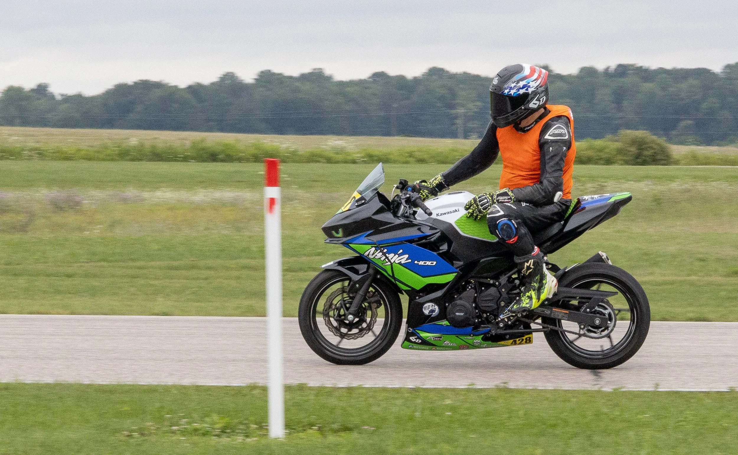 Motorcycle rider wearing a helmet with an American flag design, an orange jacket, and black riding gear, riding a Kawasaki Ninja 400 on a road with a grassy field and trees in the background.