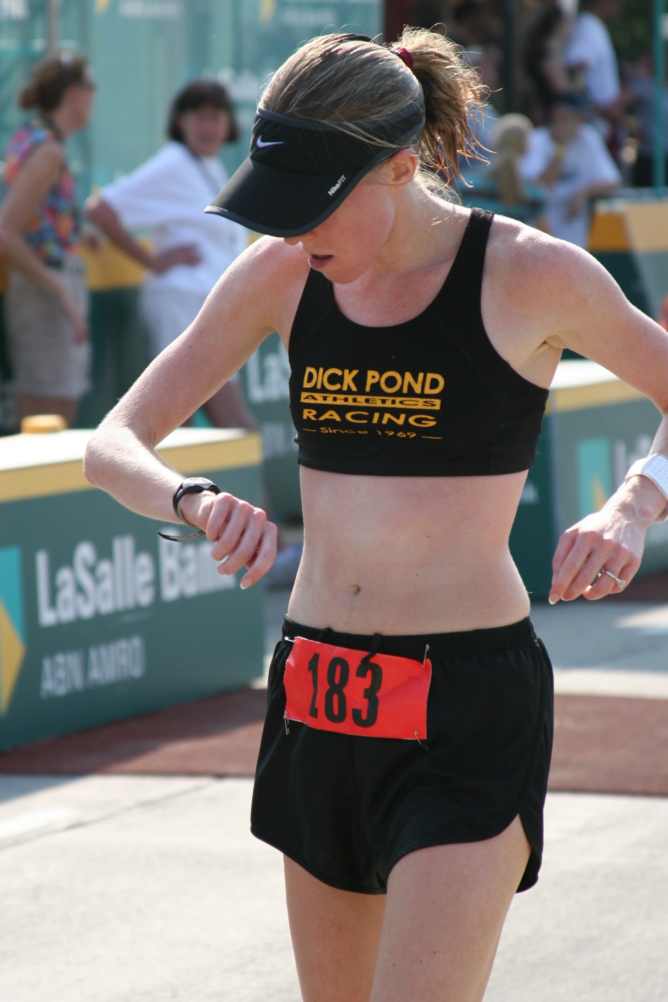 Female runner wearing black Nike sports visor, black sports bra with yellow text, black shorts, red bib number 183, and a black wristwatch, looking down during a race with spectators in the background.
