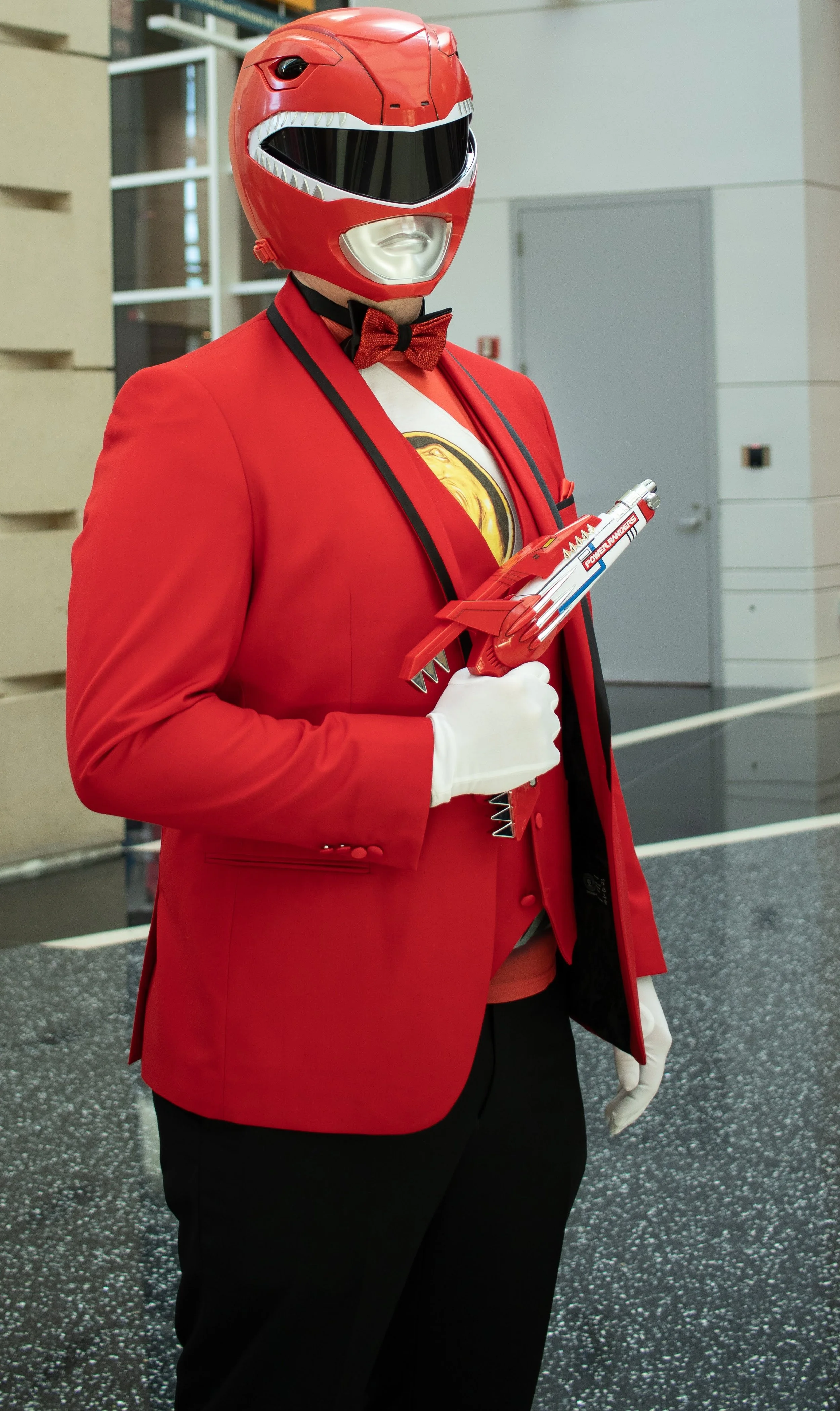 Person dressed as the Red Power Ranger, wearing a red tuxedo jacket, black pants, red bow tie, white gloves, and a red Power Ranger helmet, holding a red toy gun.