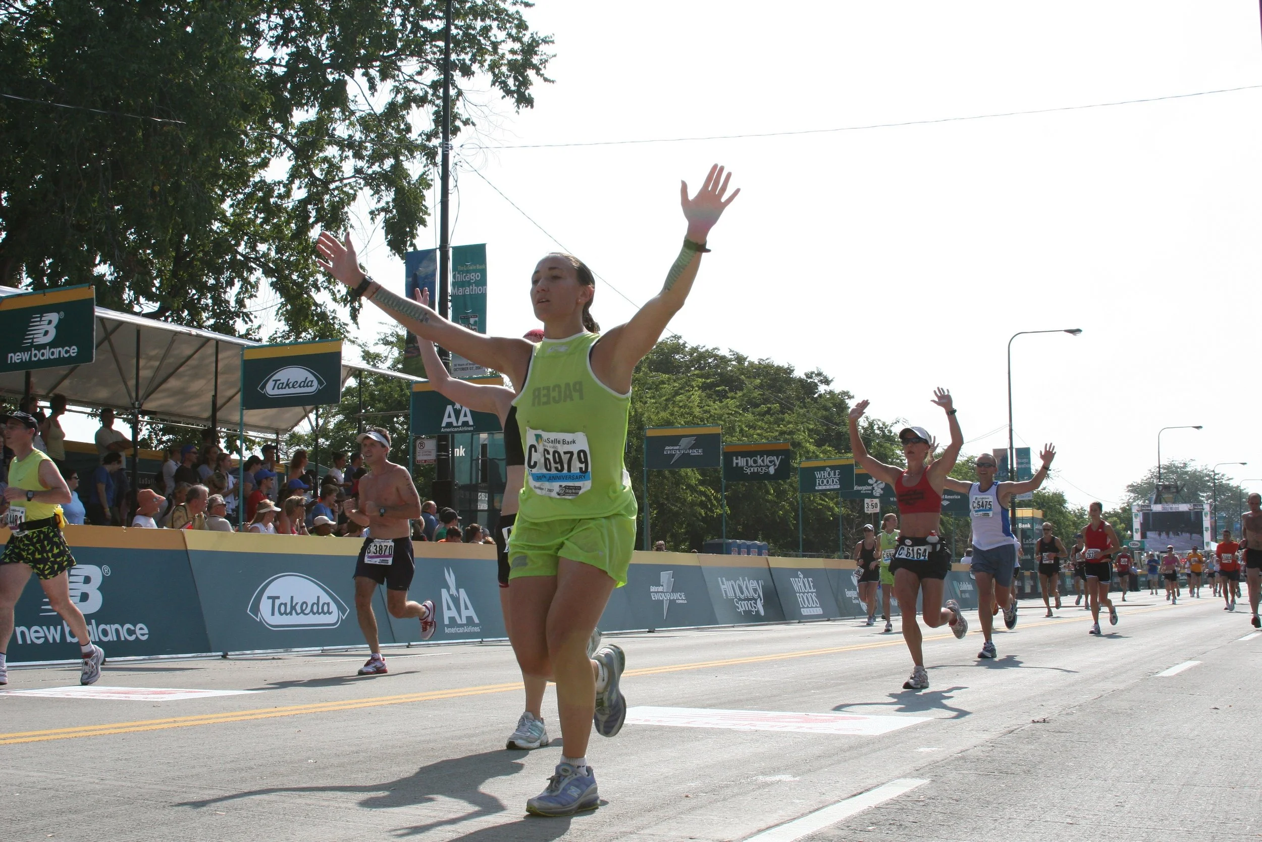A woman running in a marathon with her arms raised, surrounded by other runners and spectators, during daytime near a finish line with banners and trees in the background.
