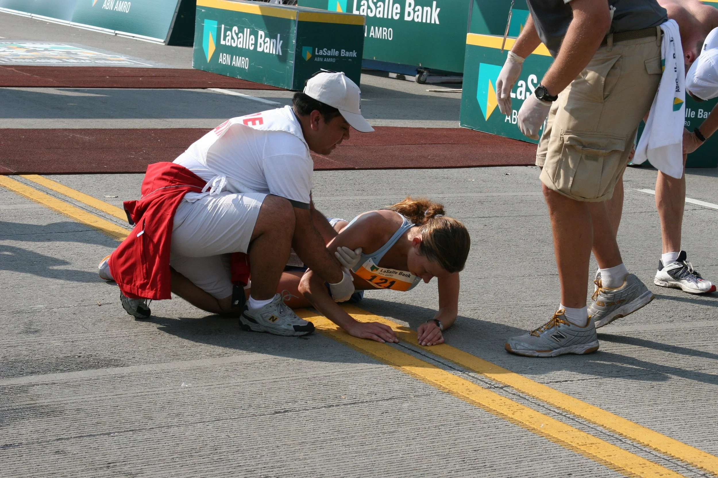 A woman has collapsed at a running race finish line, with a man and officials attending to her. The woman is on her hands and knees, and the man is crouched next to her, checking on her. Two officials stand nearby. The event is sponsored by LaSalle B