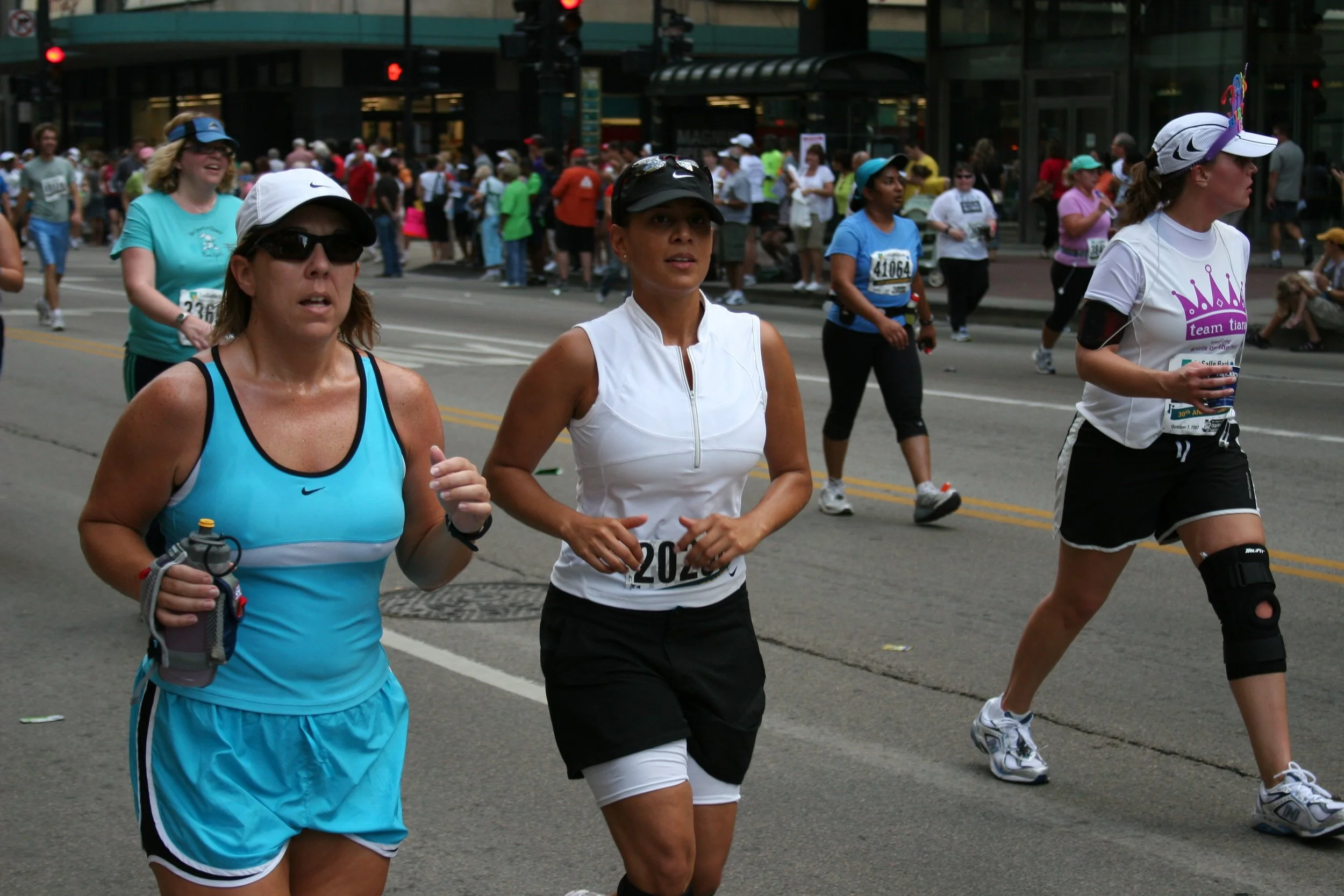 A group of women running in a marathon event on a city street, with many spectators and onlookers in the background.
