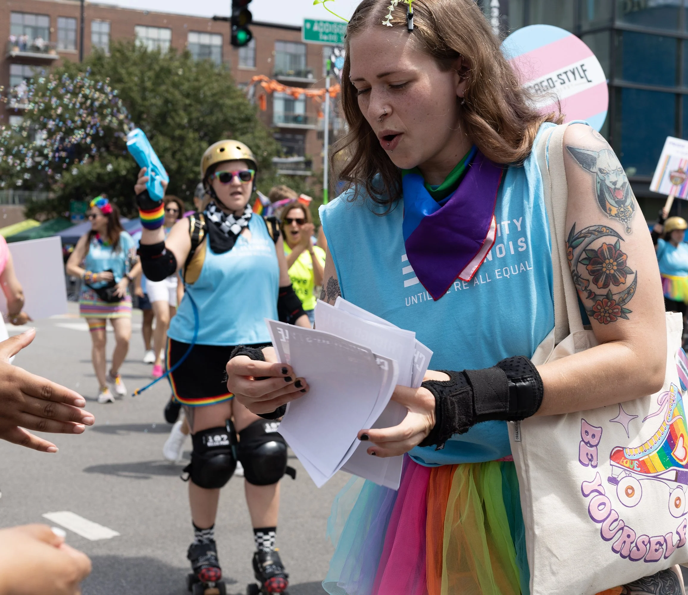A person with tattoos reading a paper at a pride event, dressed in rainbow-colored clothing, surrounded by other participants, some on roller skates, with a rainbow flag banner and festive decorations in the background.