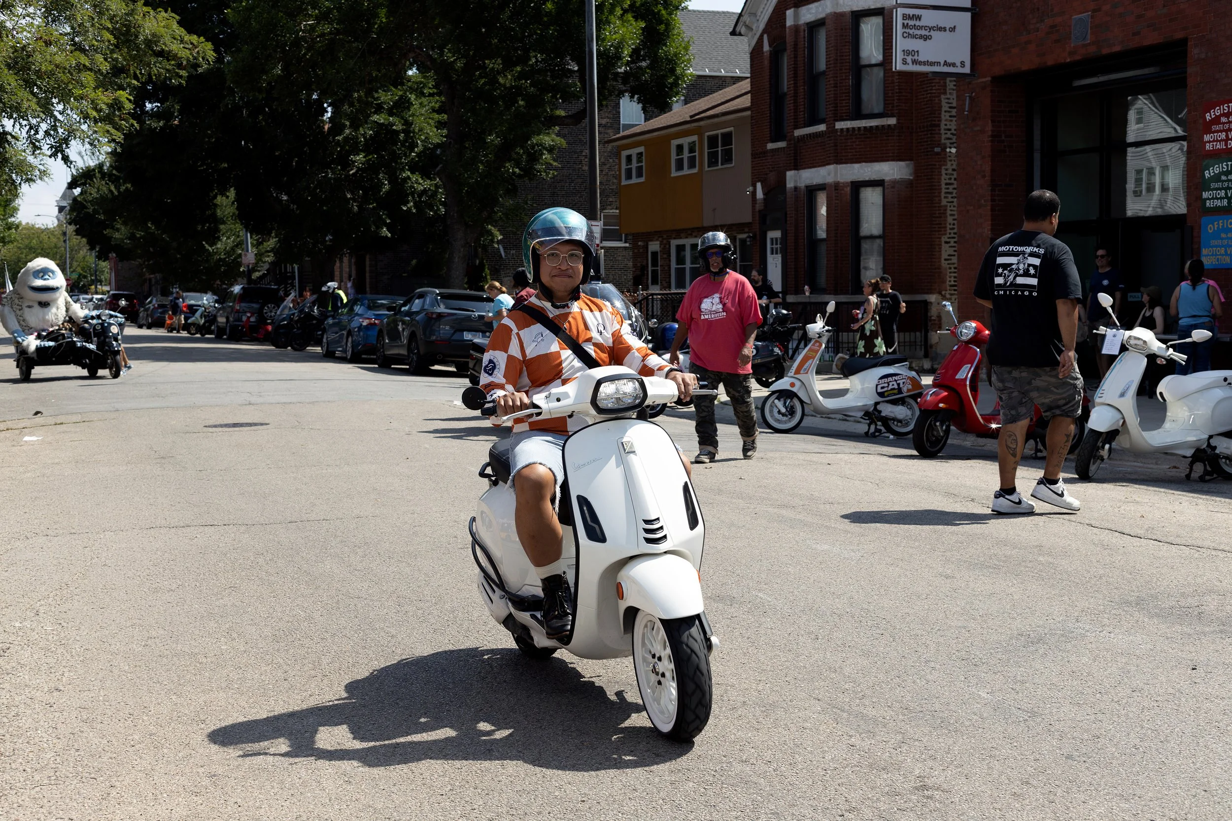 A person riding a white Vespa scooter on a city street, wearing a helmet and glasses. In the background, there are people, parked scooters, and cars, along with brick buildings and trees.