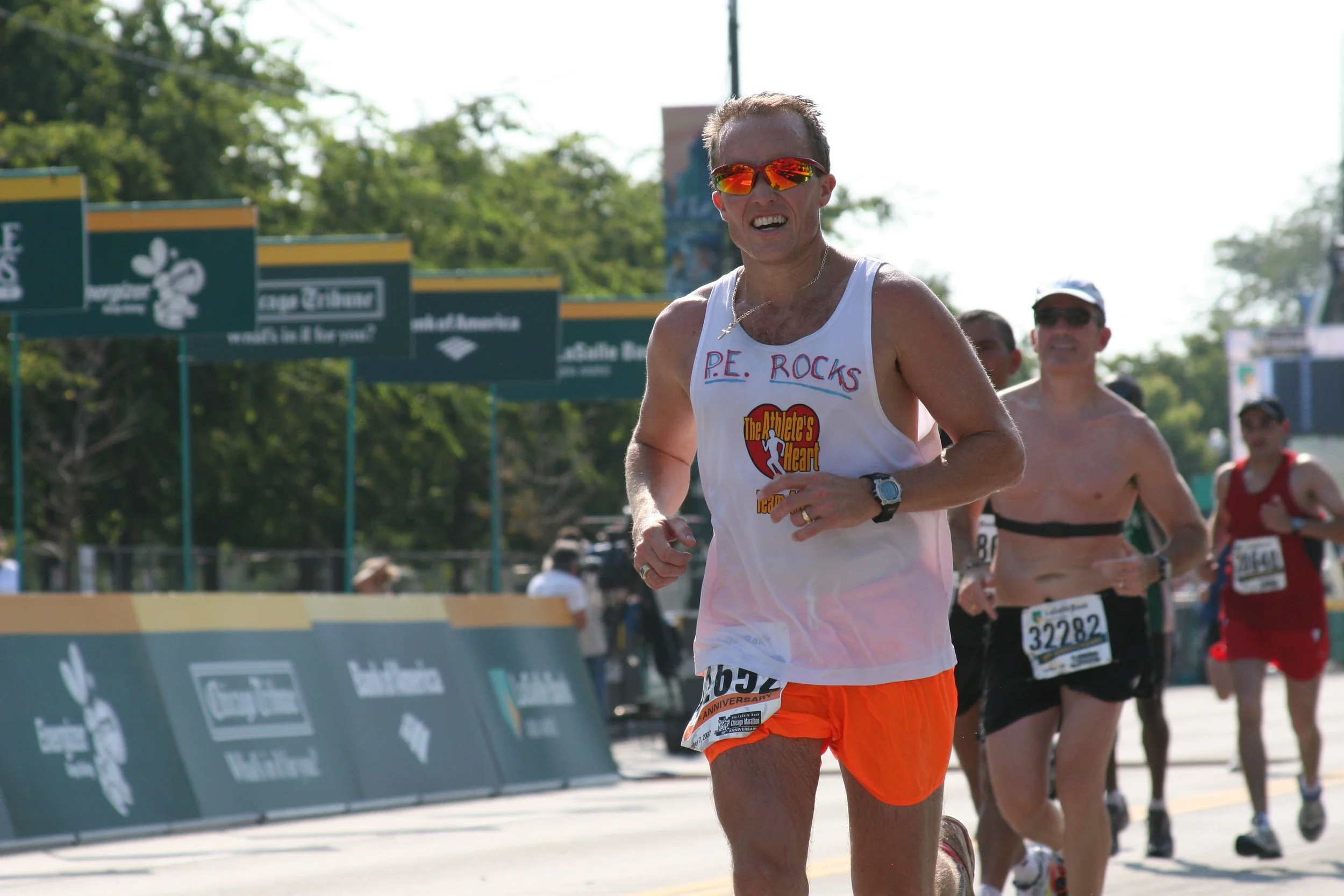 Male runner in a white tank top with "P.E. ROCKS" written on it and orange shorts, smiling and wearing sunglasses, participating in a marathon along with other runners on a sunny day.