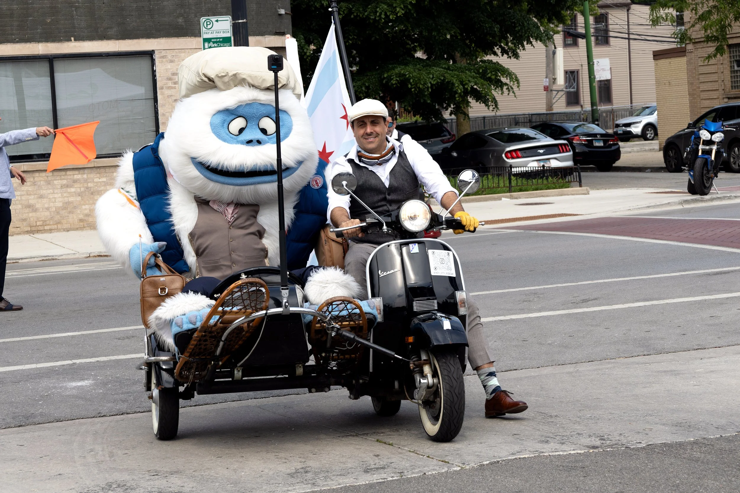 A person riding a scooter with a person in a mascot costume sitting in a sidecar. The mascot appears to be a blue and white fluffy animal or creature. The scene takes place on a city street with parked cars and buildings in the background.