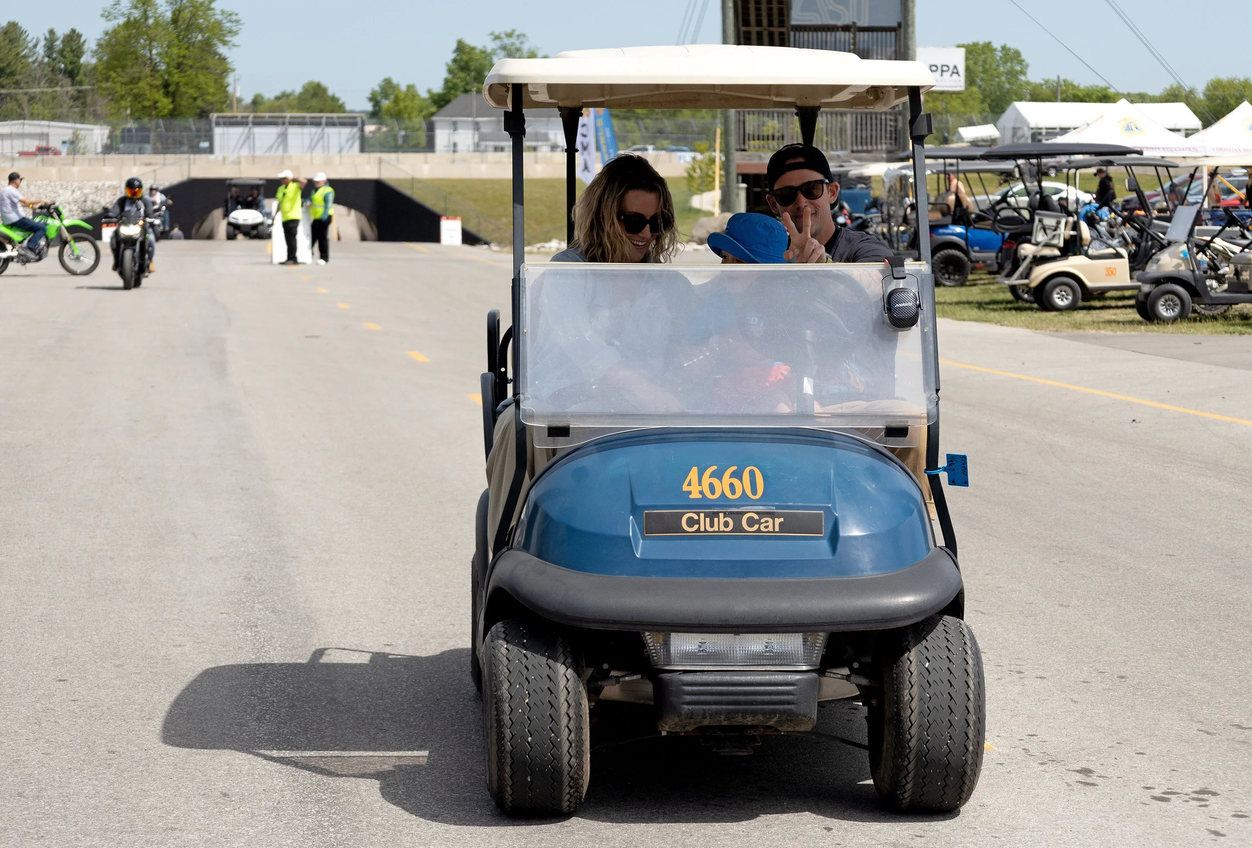 A golf cart with the number 4660 and label 'Club Car' parked on a lot. Two people are sitting inside the golf cart, smiling and making a peace sign. The background includes more golf carts, people, greenery, and a clear sky.