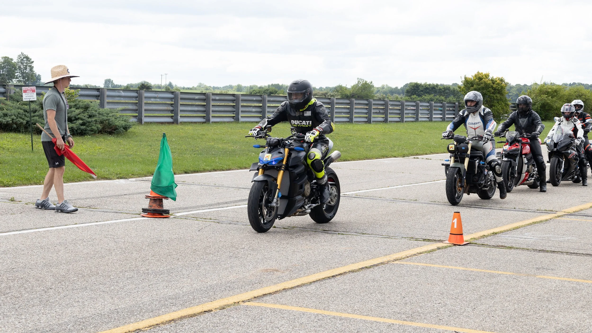 Group of motorcyclists participating in a riding event, waiting in line with a coordinator holding a red flag at a designated checkpoint on a cloudy day.
