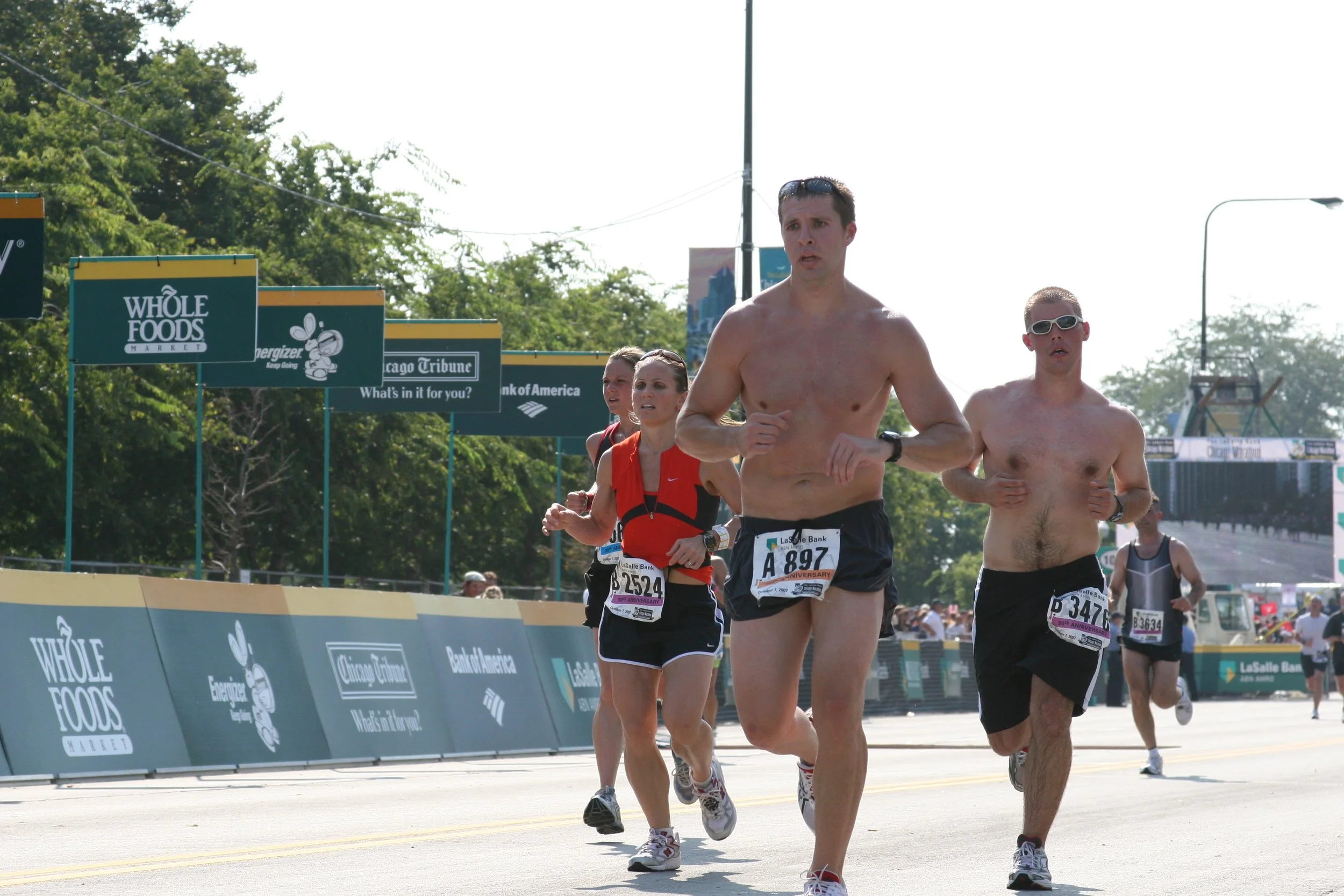 Group of marathon runners participating in a race on a sunny day, running along a city street with advertisements for Whole Foods Market and other sponsors.