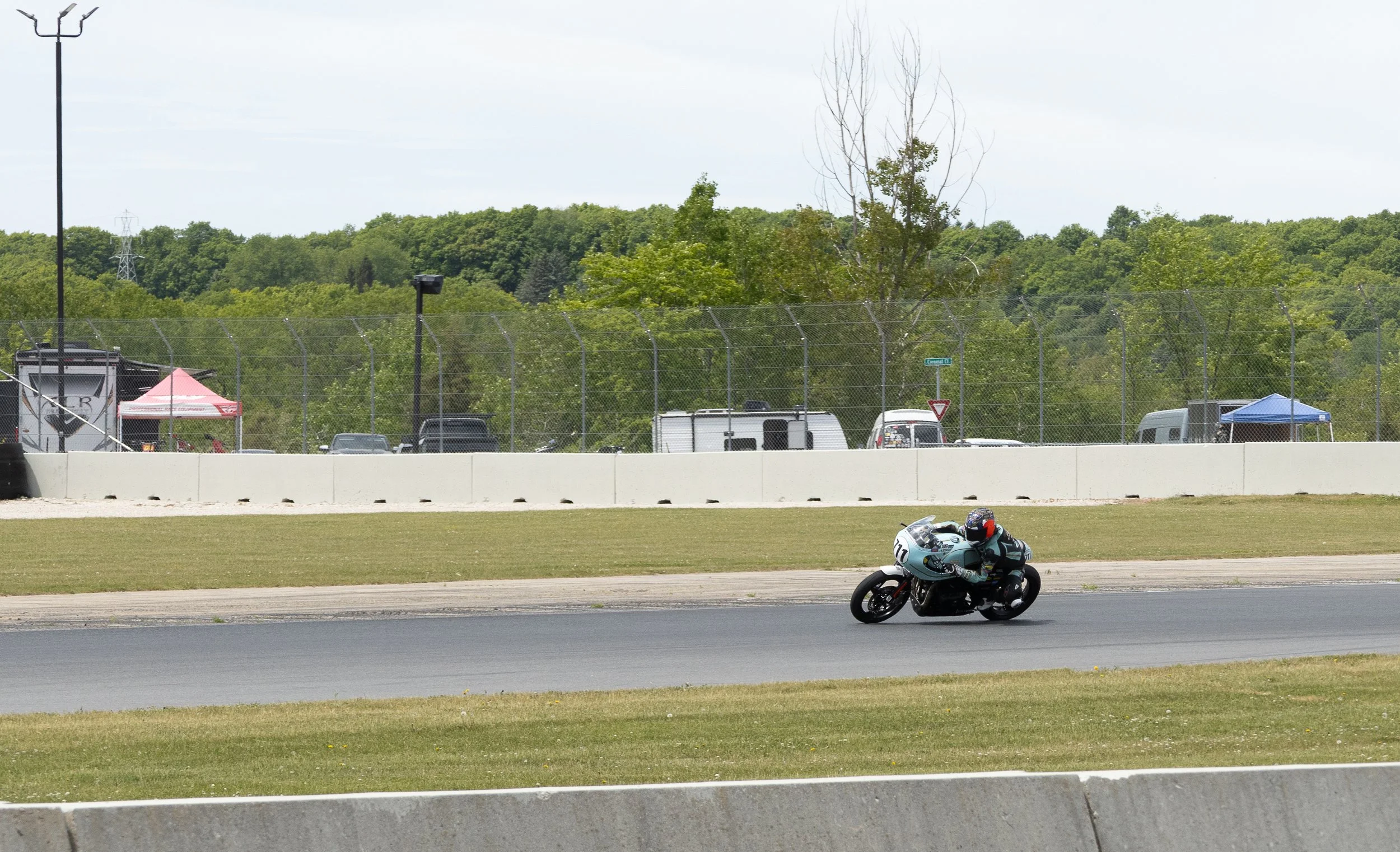 A motorcycle racing on a track with safety barriers, tents, and a parking lot with cars in the background.