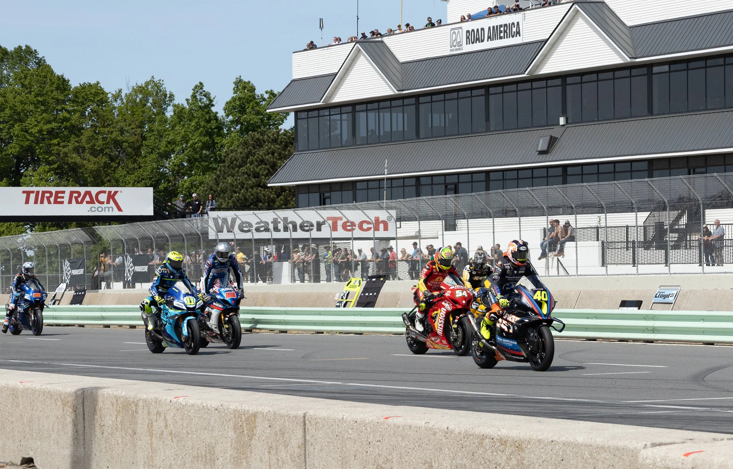 Motorcycle racers in colorful gear racing on a track with spectators watching from a tall building and banners advertising Tire Rack and WeatherTech.