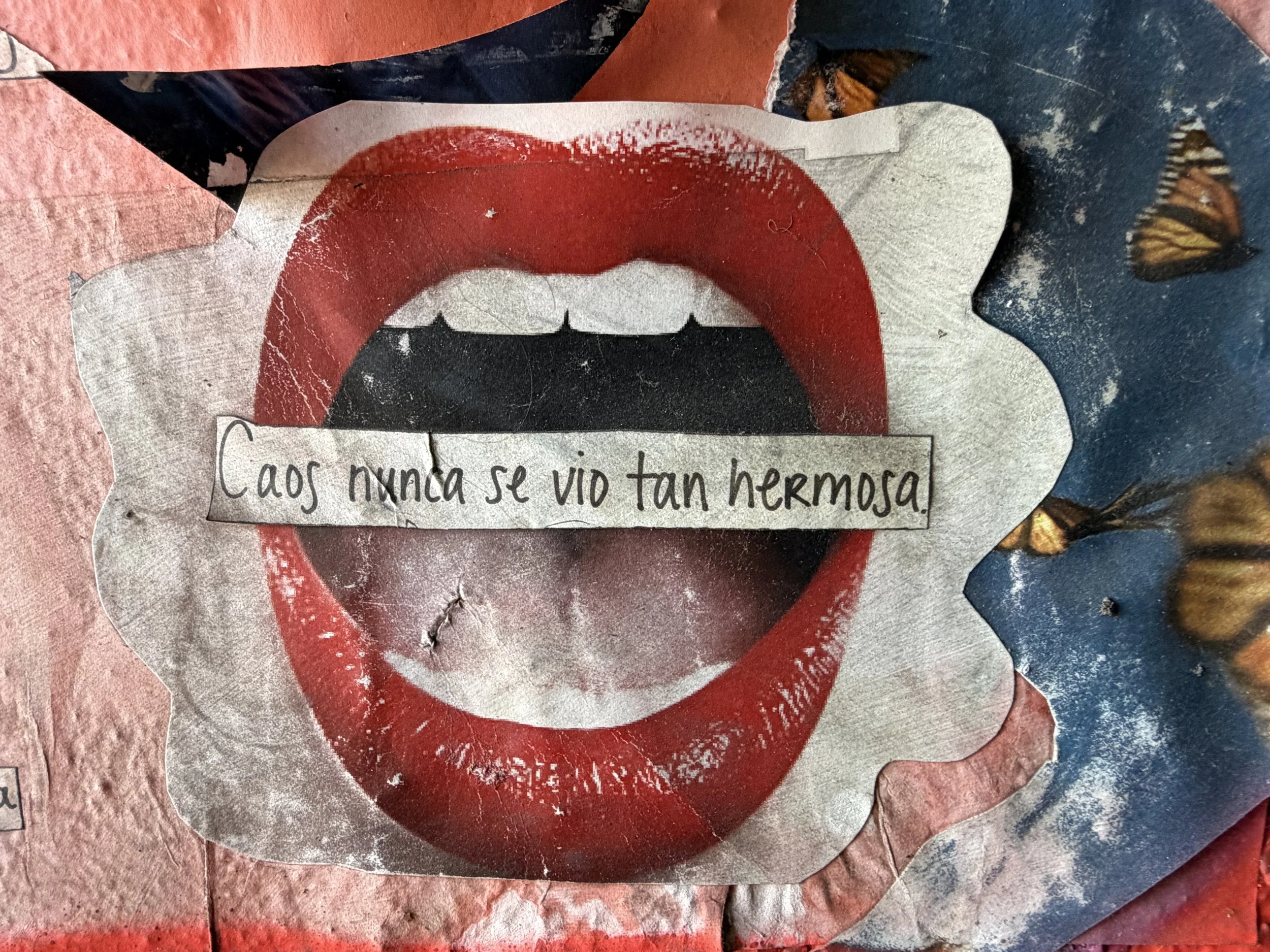 A torn piece of paper with a printed image of lips and a partial quote in Spanish that reads 'Cao nunca se vio tan hermosa.' The paper is laid on a pink and blue textured surface next to a butterfly-patterned fabric.