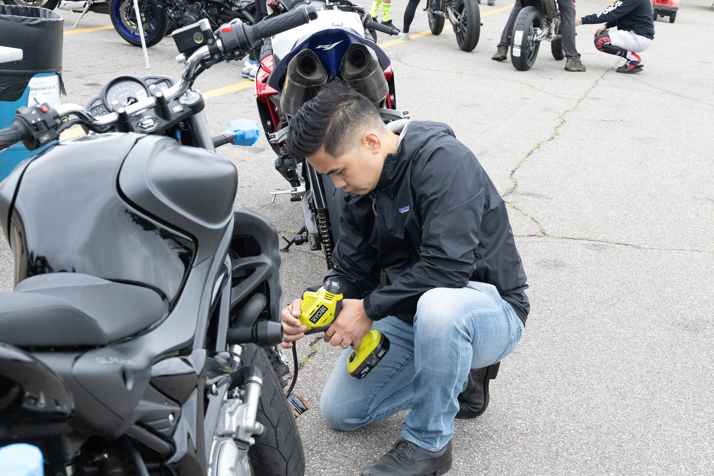 A man kneels on the ground, working on a motorcycle with power tools, in an outdoor parking lot. Several other motorcycles and people are visible in the background.