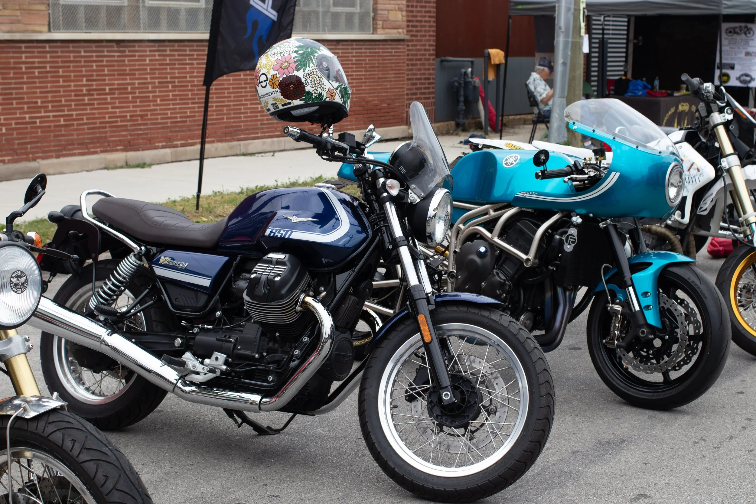 A lineup of motorcycles parked on a city street during a sunny day, with brick buildings and pedestrians in the background.