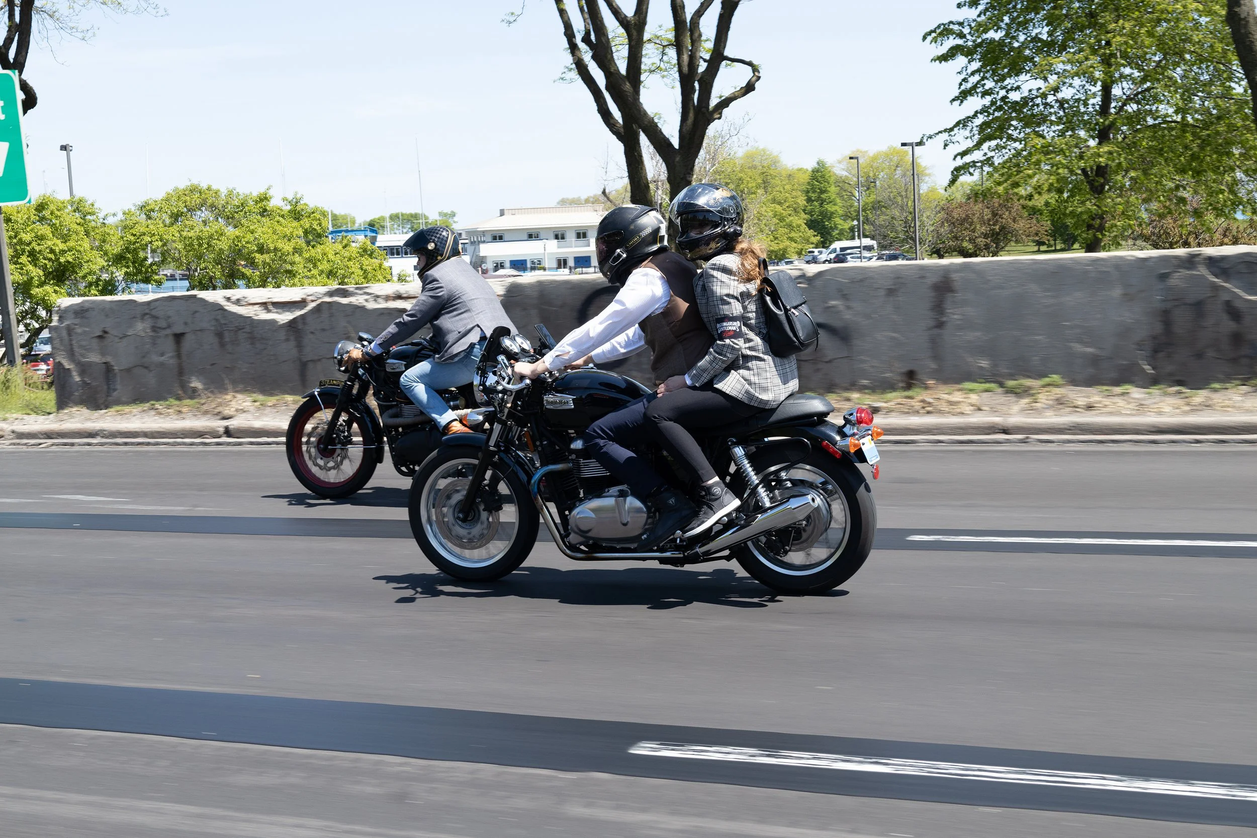 Three people riding motorcycles on a road, all wearing helmets. One is a woman with long hair, dressed in a plaid blazer and black pants. Another is a man in a suit. The third rider is another man dressed casually. The background includes trees and p