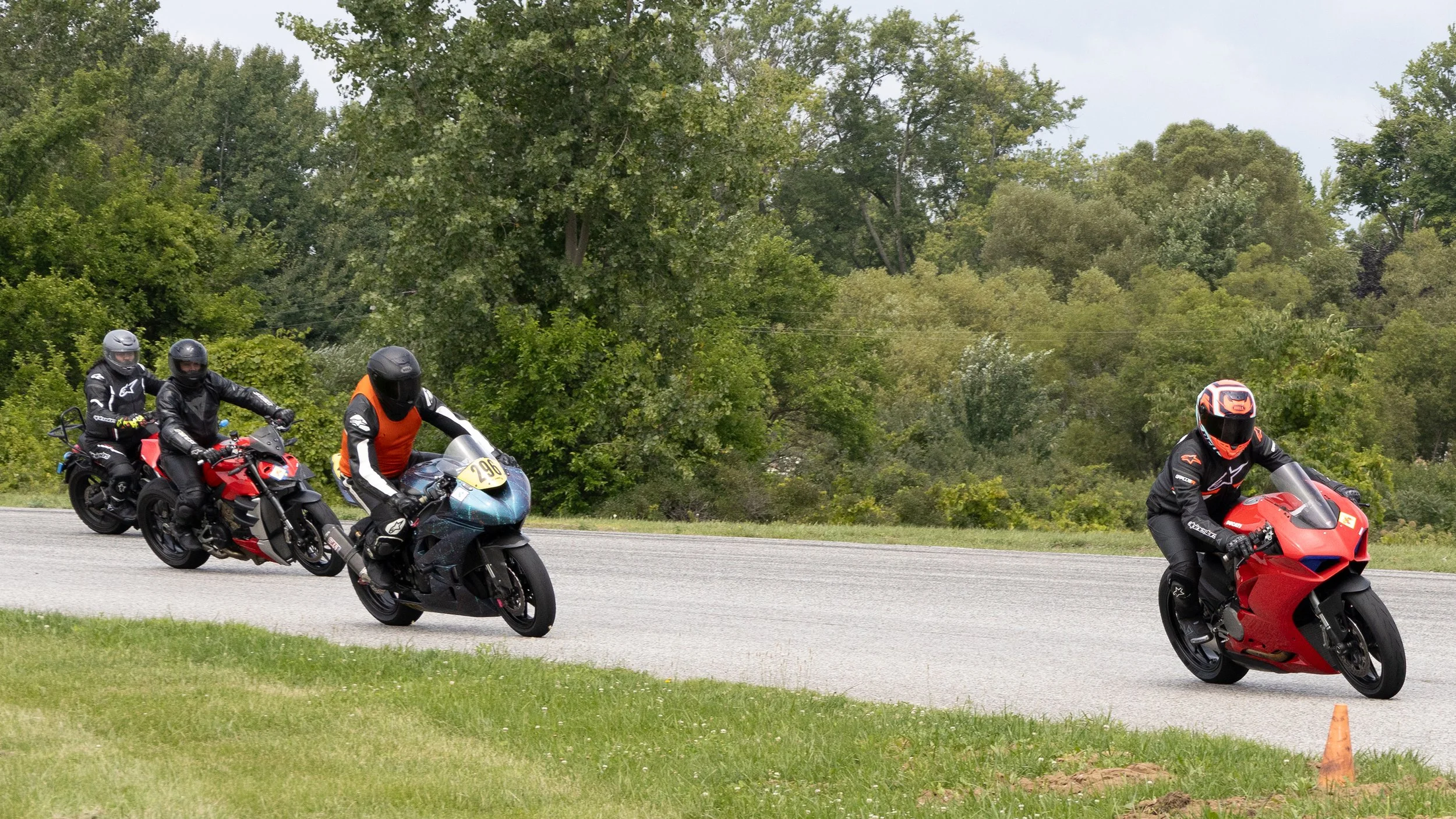 Four motorcyclists wearing protective gear, riding sport bikes on a paved track with green trees in the background.