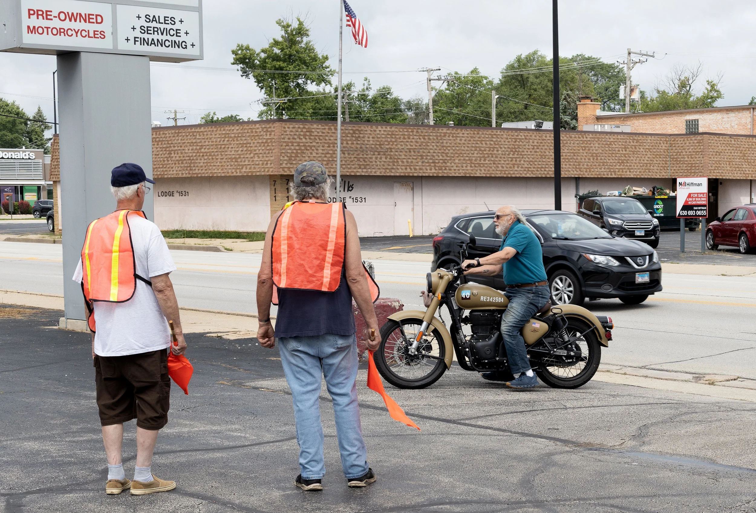 Three elderly men stand on the sidewalk near a woman wearing a safety vest, with a man on a motorcycle nearby. They are in front of a commercial building with a sign advertising pre-owned motorcycles and related services. An American flag is visible 