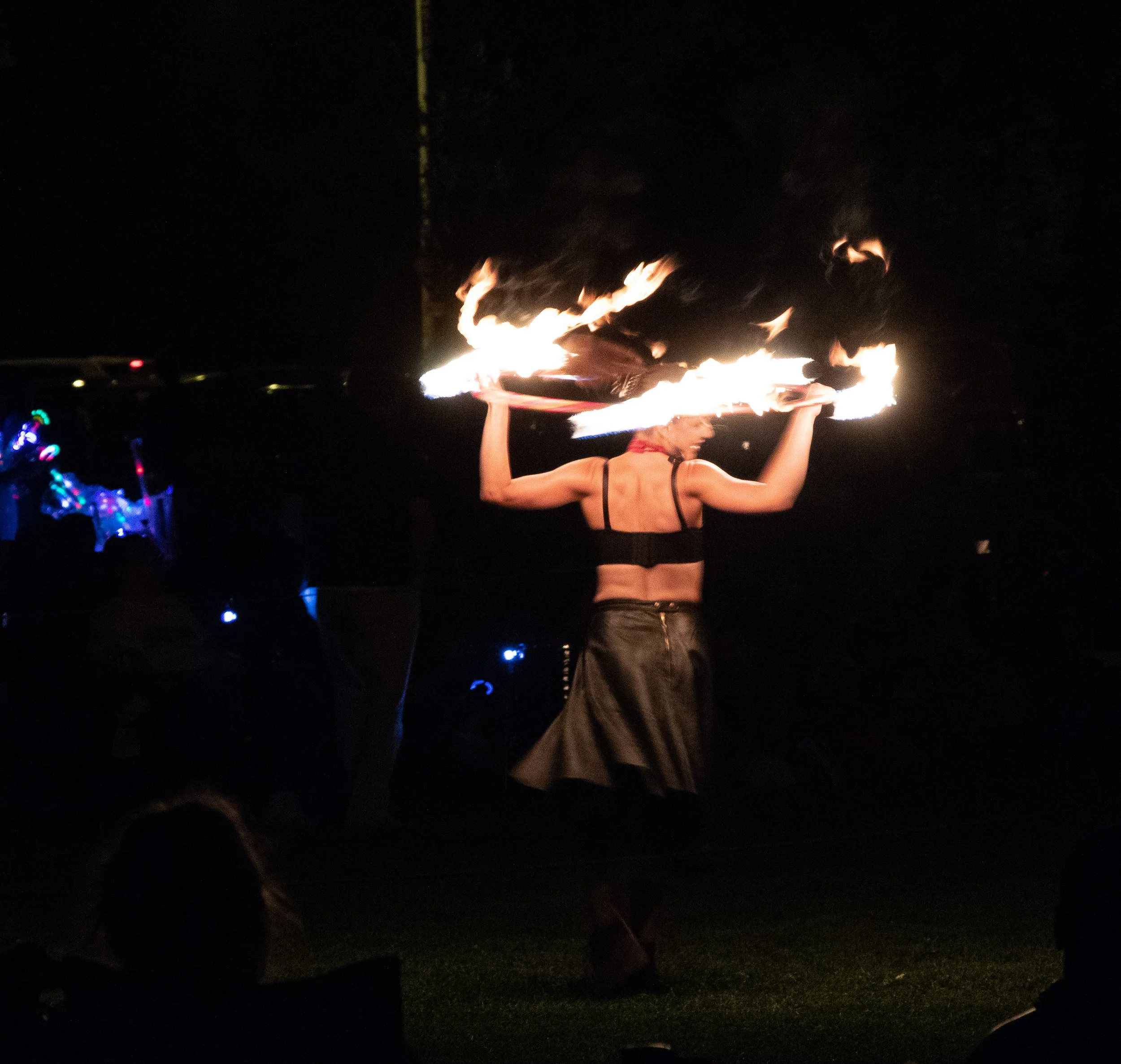 A woman holding a flaming tray at night, with dark surroundings and some colorful lights visible in the background.