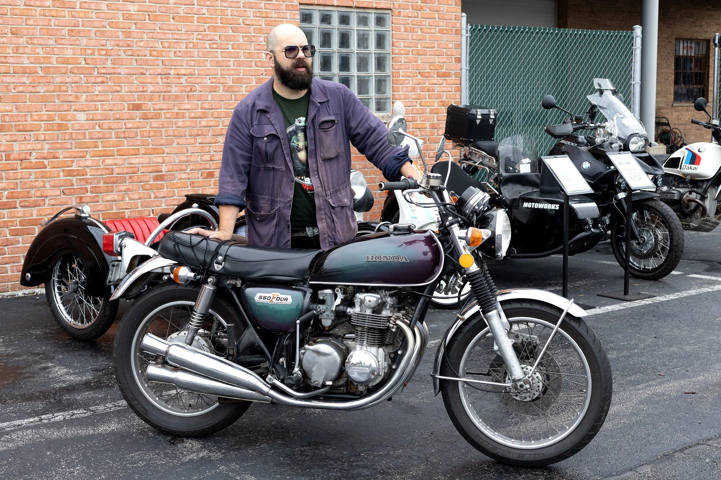 A man with a shaved head, beard, and sunglasses stands beside a vintage Honda motorcycle, with several other motorcycles displayed nearby on a blacktop lot against a brick wall.