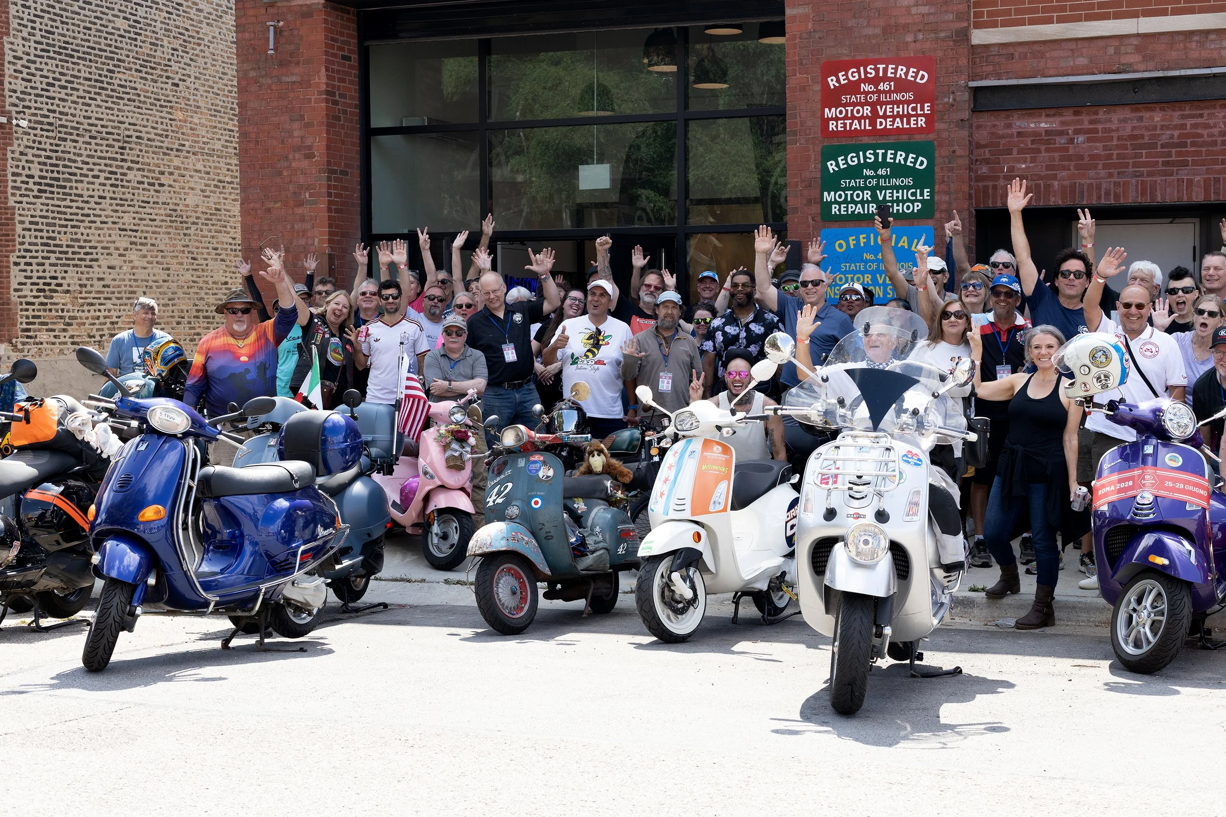 Group of people smiling and waving in front of a building with signs indicating motor vehicle registration, repair shop, and dealership, standing behind a lineup of various scooters and motorcycles.