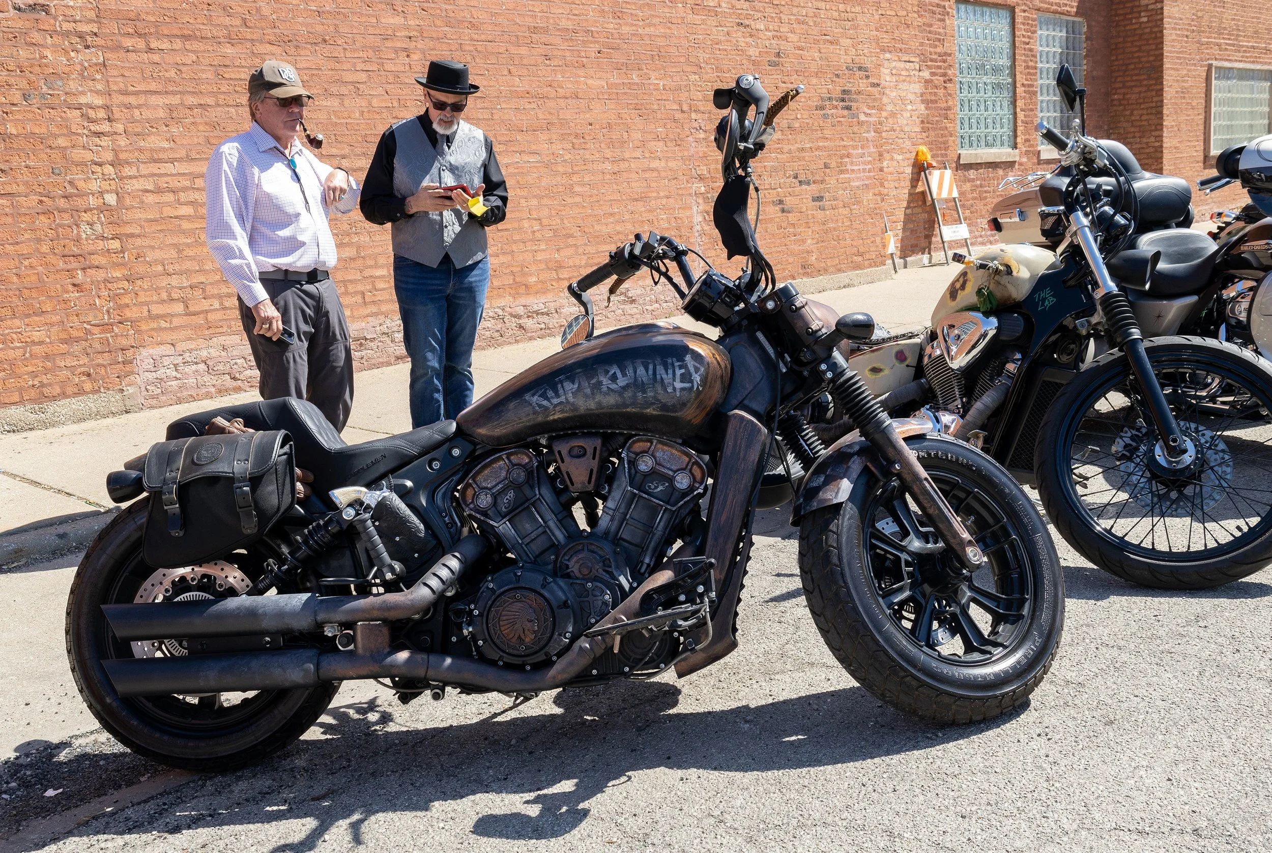 Two men standing beside motorcycles against a red brick wall. One man is smoking a pipe, wearing sunglasses and a hat, while the other is using a smartphone, wearing a hat and sunglasses.