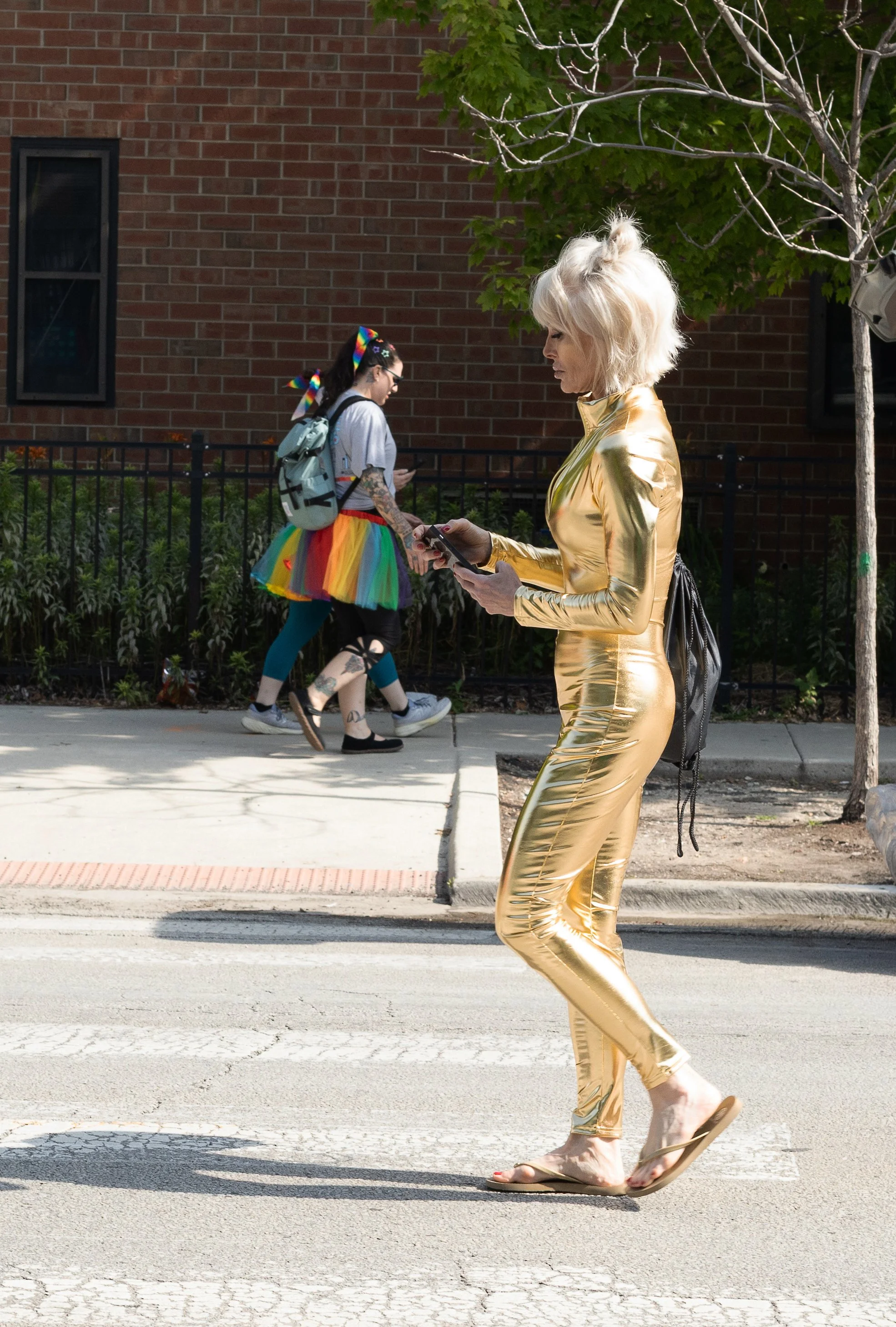 A woman in a shiny gold jumpsuit and sandals walking across a crosswalk, looking at her phone, with two women in colorful outfits walking behind her.