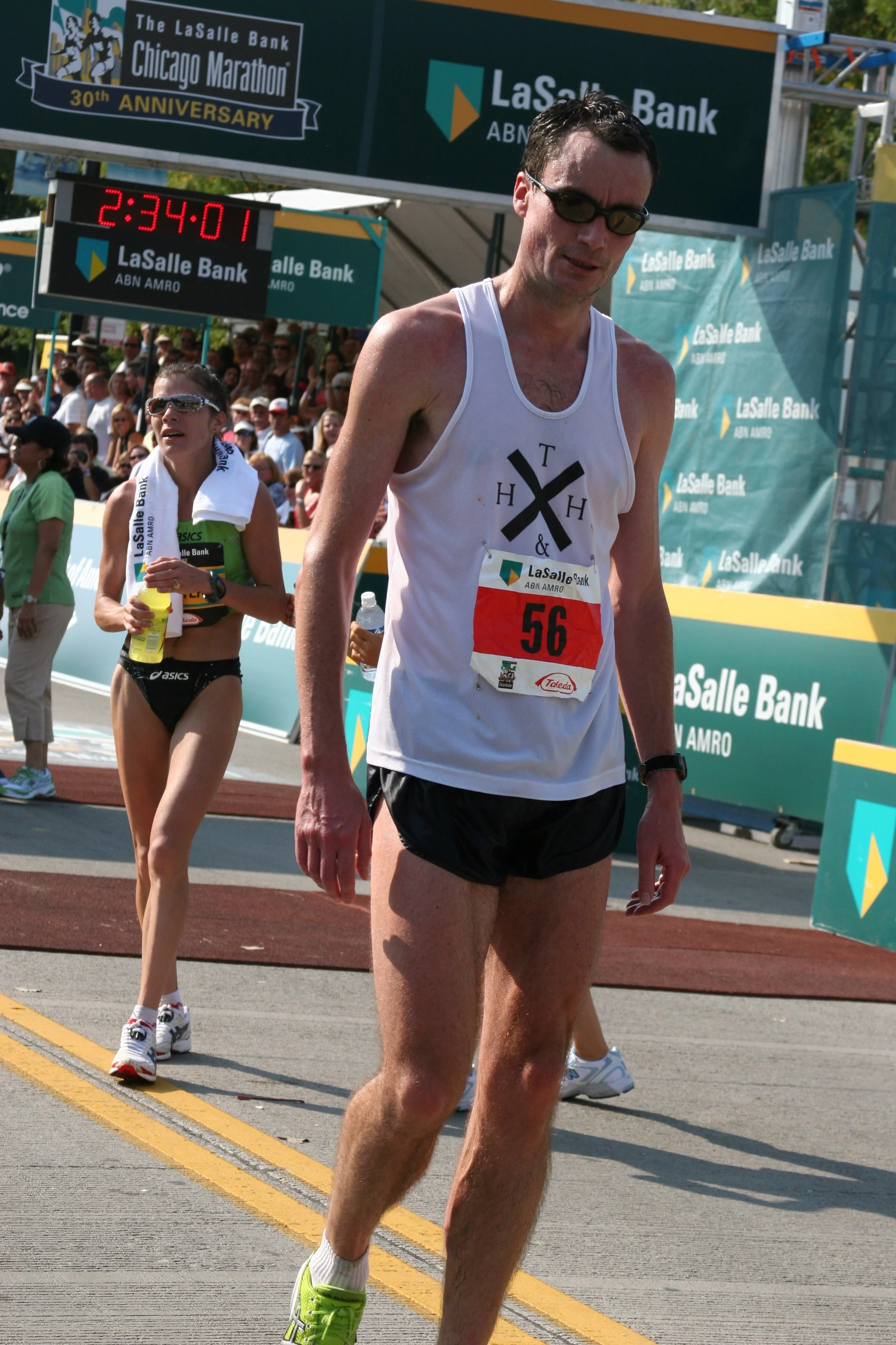 Male marathon runner in white tank top with the number 56, wearing black shorts and sunglasses, running in a race with a female runner in black shorts and a green top behind him, crowd in the background, and a banner with LaSalle Bank and Chicago Mar