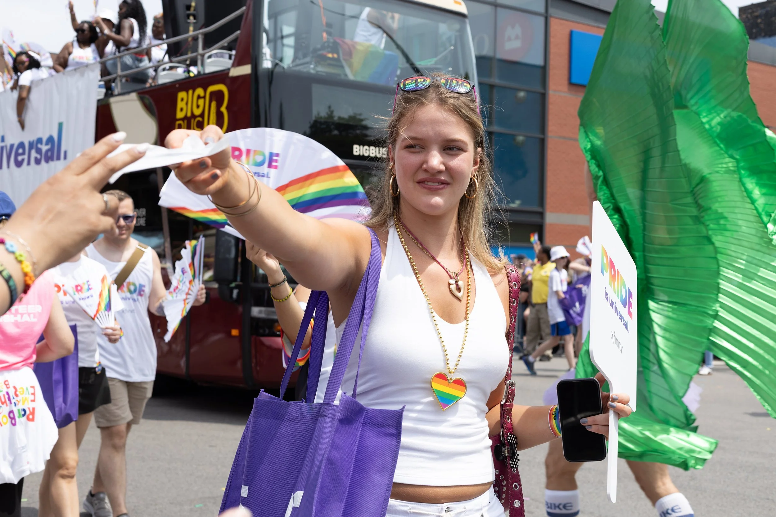 Young woman participating in a pride parade, wearing rainbow-themed accessories and holding a sign, with parade floats and other participants in the background.
