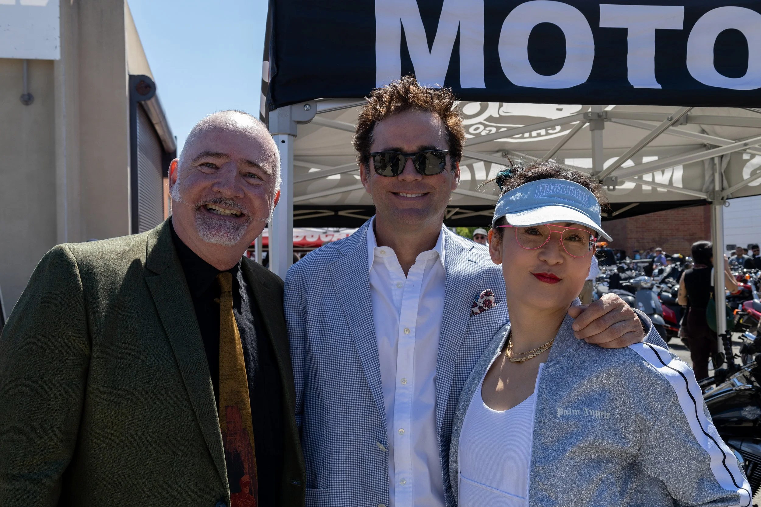 Three smiling people posing outdoors at a daytime event, with a tent and motorcycles in the background.